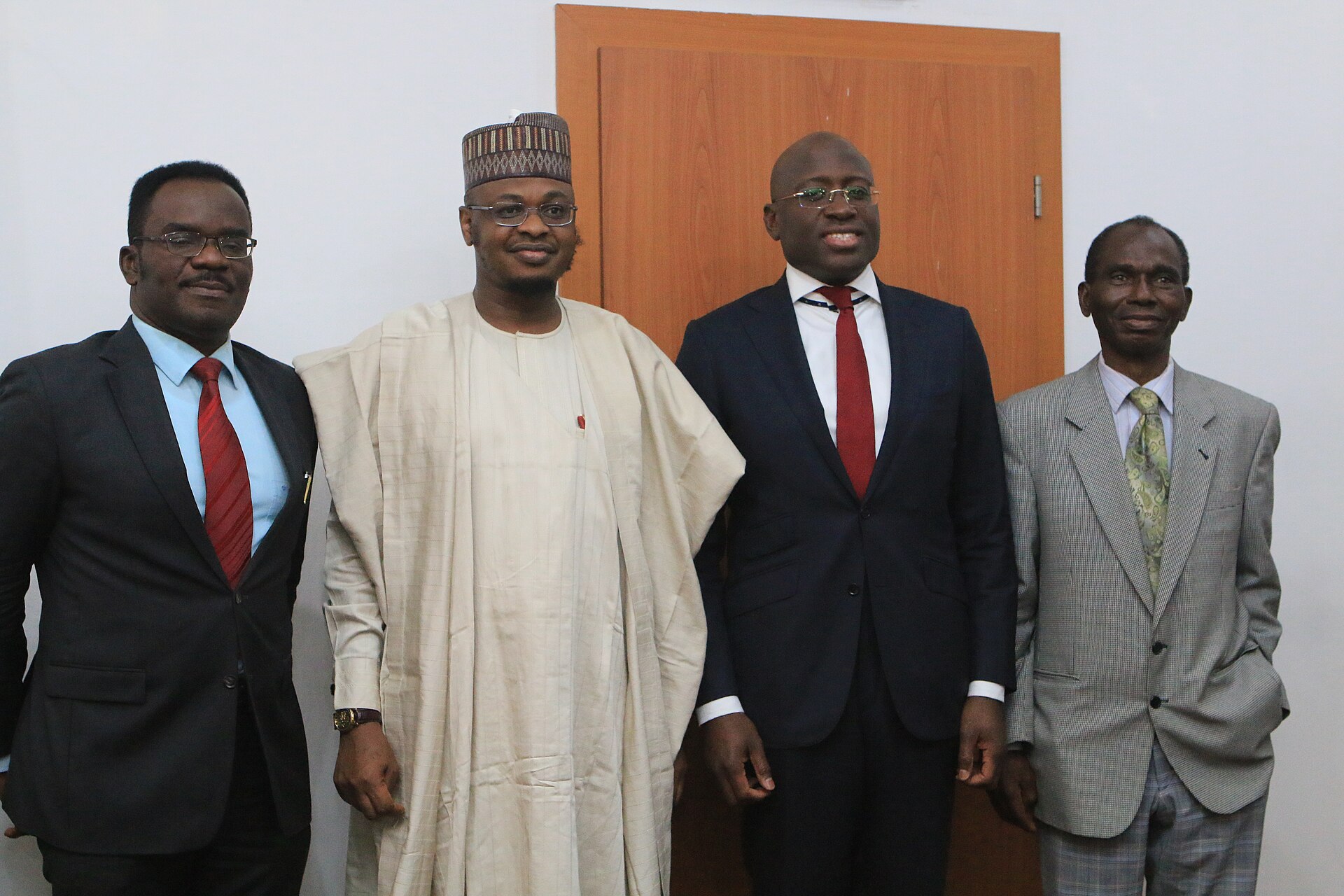 Four men standing in front of a wooden door; the second from left wears a cream kaftan and an embroidered cap, the others wear suits