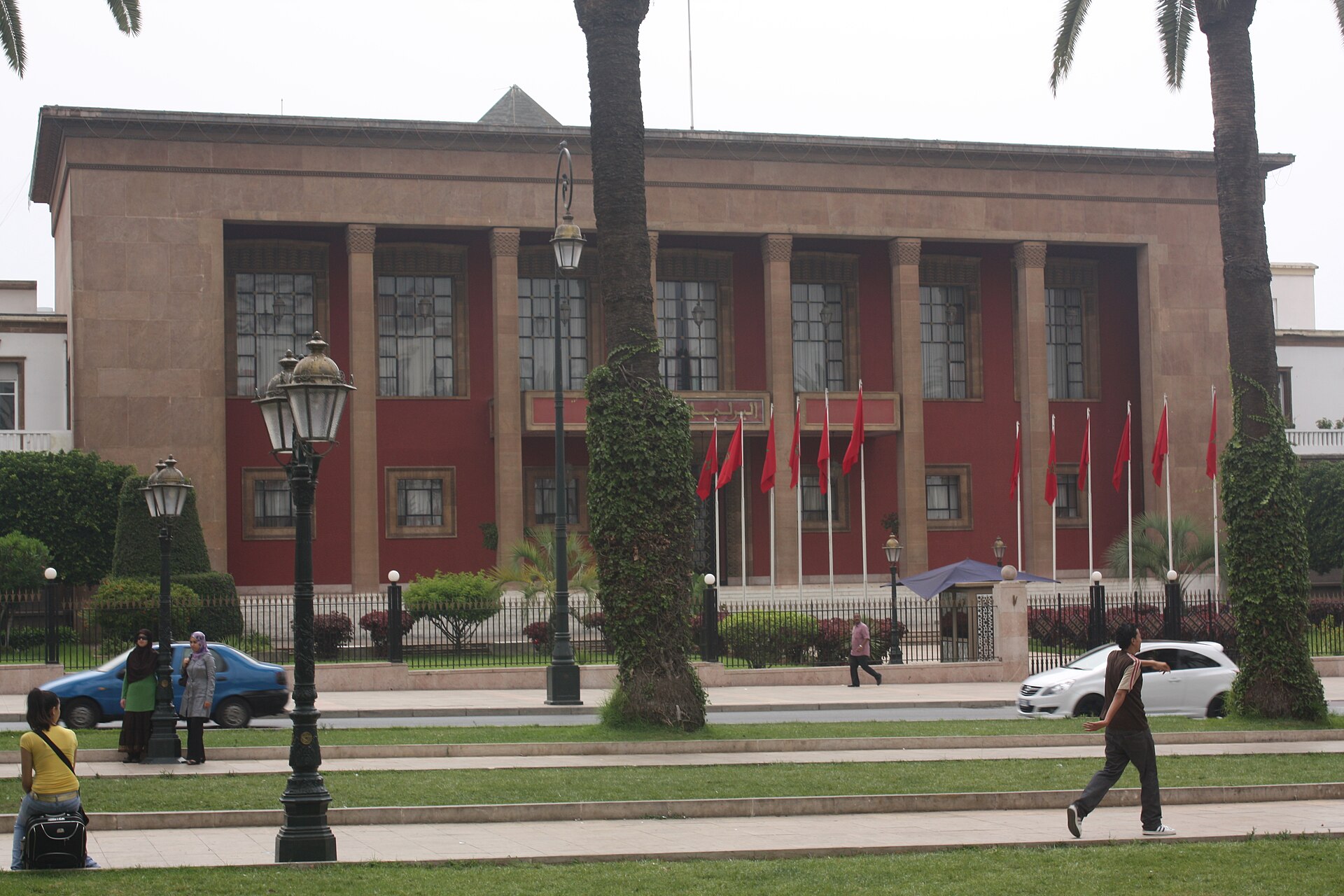 The red and sandstone facade of the Moroccan Parliament in Rabat behind palm trees, street lamps and pedestrians