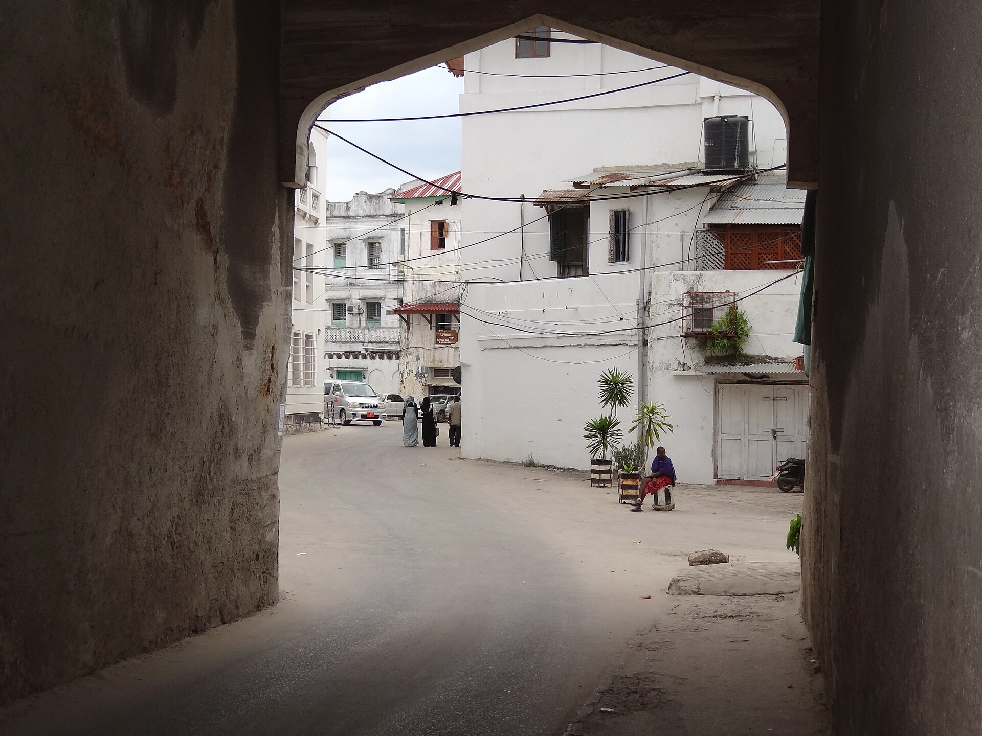 A whitewashed street in Stone Town seen through a dark arched passage, with a few people and parked cars