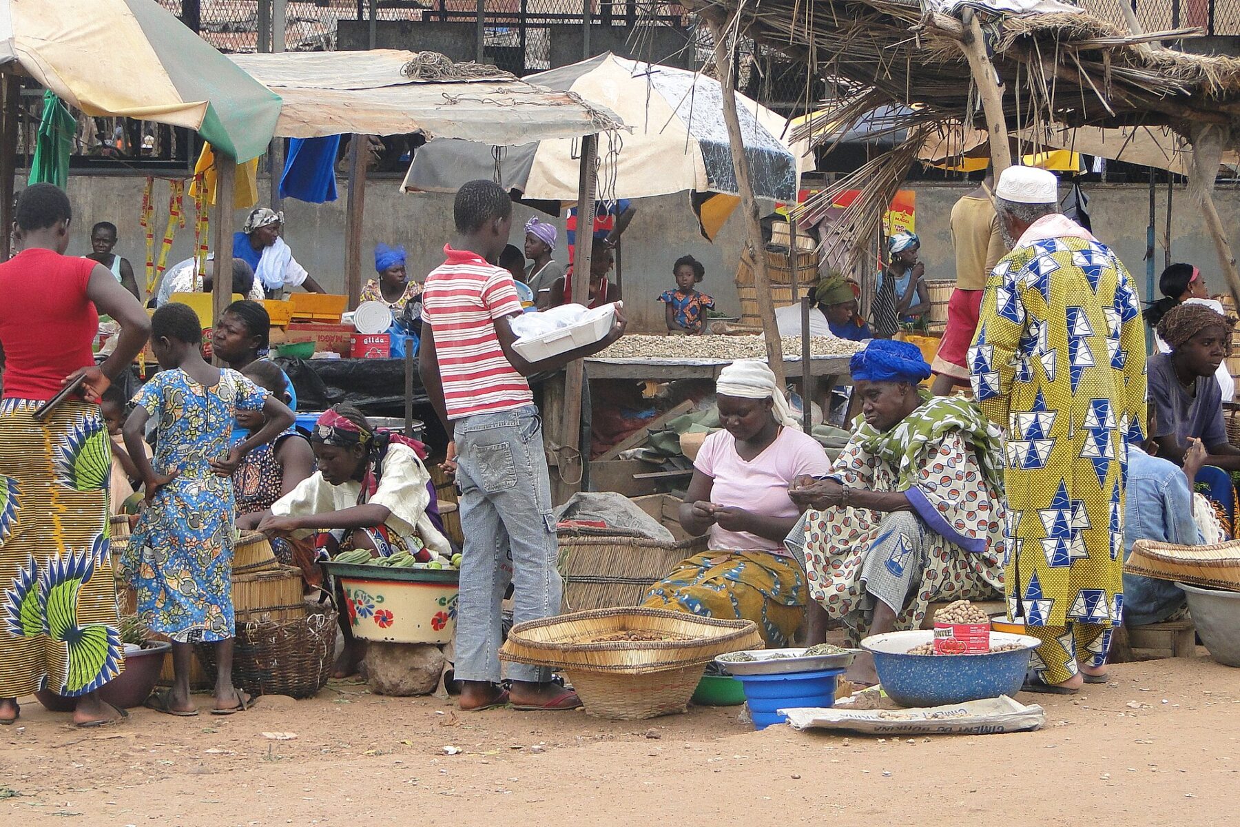 Women selling grain and produce from baskets and bowls under shade awnings at an open-air market in Gaoua, Burkina Faso