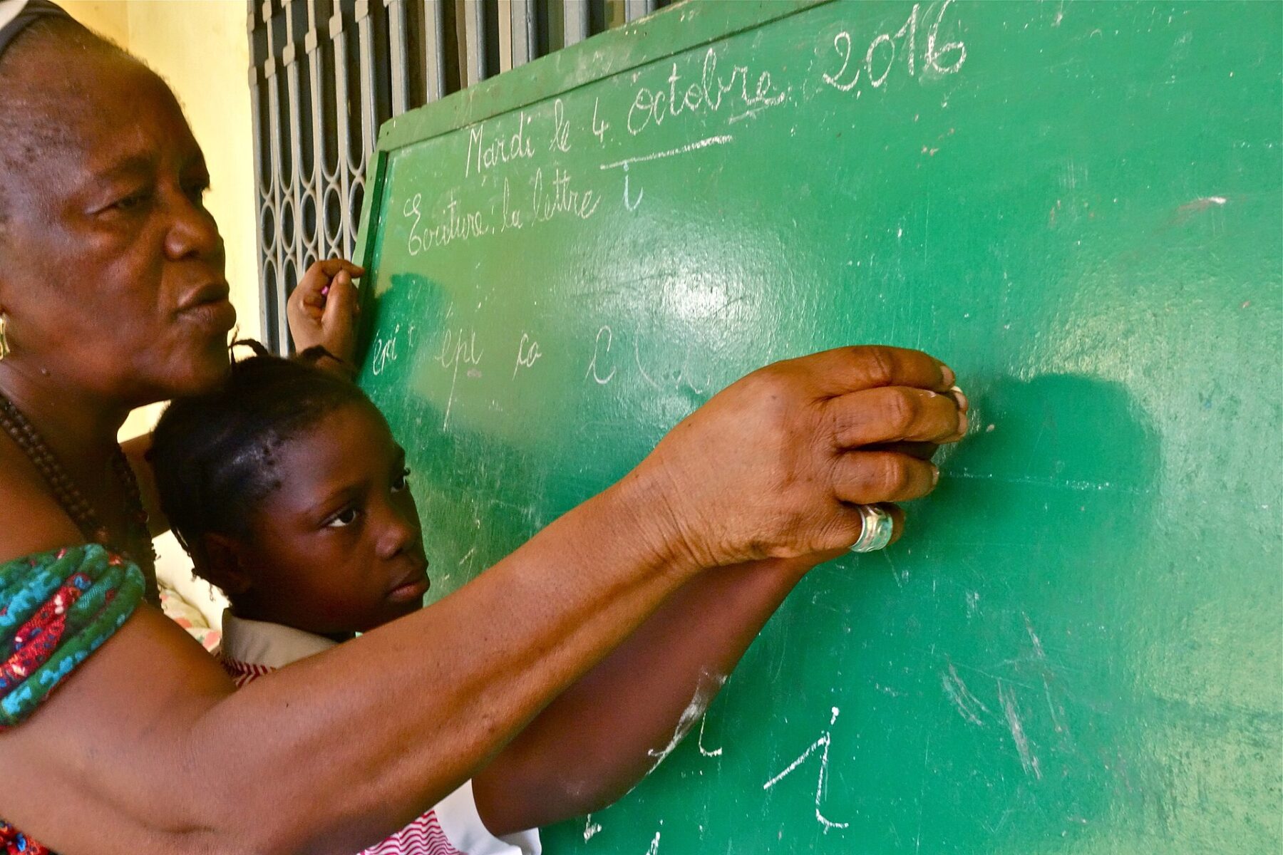 A teacher guides a young pupil writing letters on a green chalkboard in a Guinean classroom