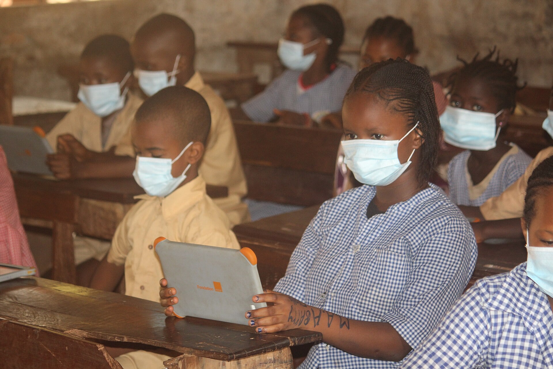 Masked pupils in school uniforms sit at wooden desks holding tablets in a classroom