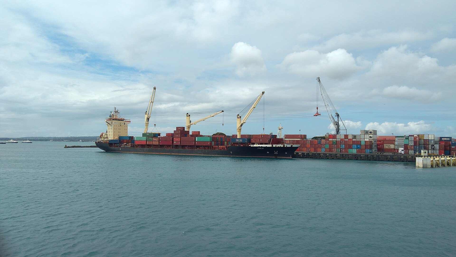 A container ship unloading at a Tanzanian quay with cranes and stacked containers under a cloudy sky