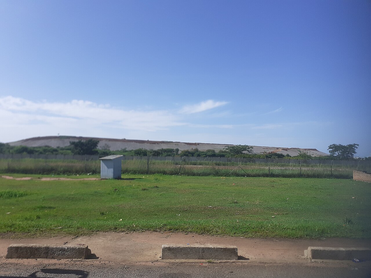 Grey waste-rock dump of the Buzwagi gold mine behind a fence and a grass field, seen from a roadside in Kahama
