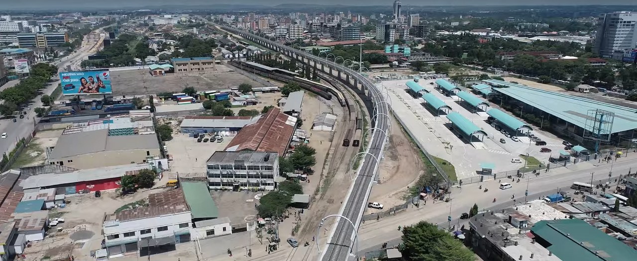 Aerial view of an elevated railway curving past an older rail yard with freight wagons and a station with teal roofs in Dar es Salaam