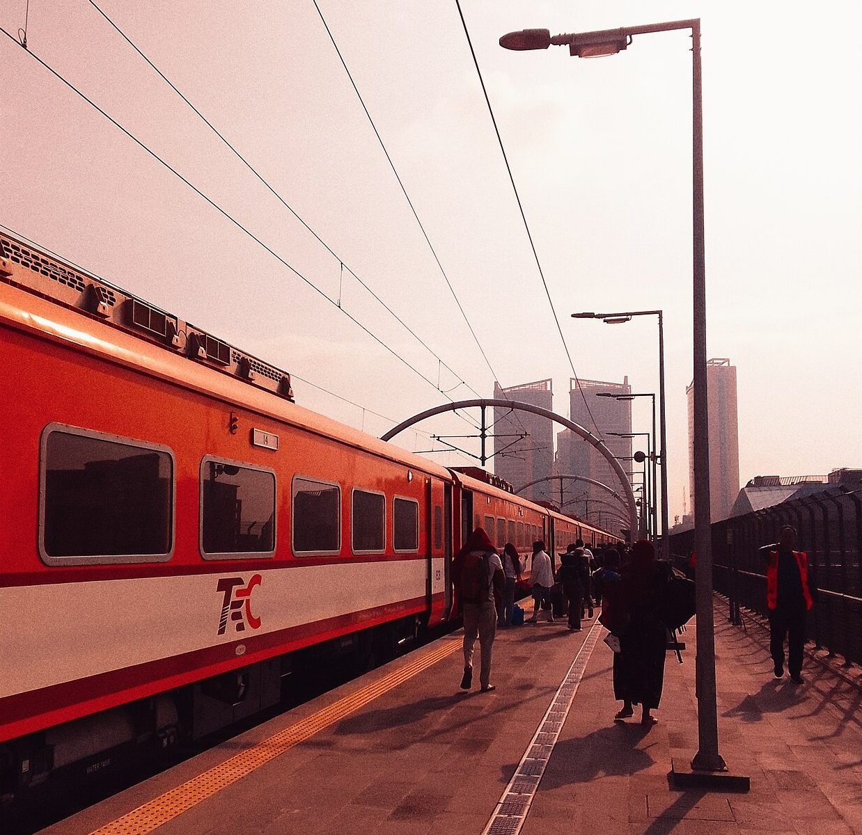 Passengers with bags walk along a platform beside a red-and-white TRC passenger train under overhead power lines, with tower blocks behind