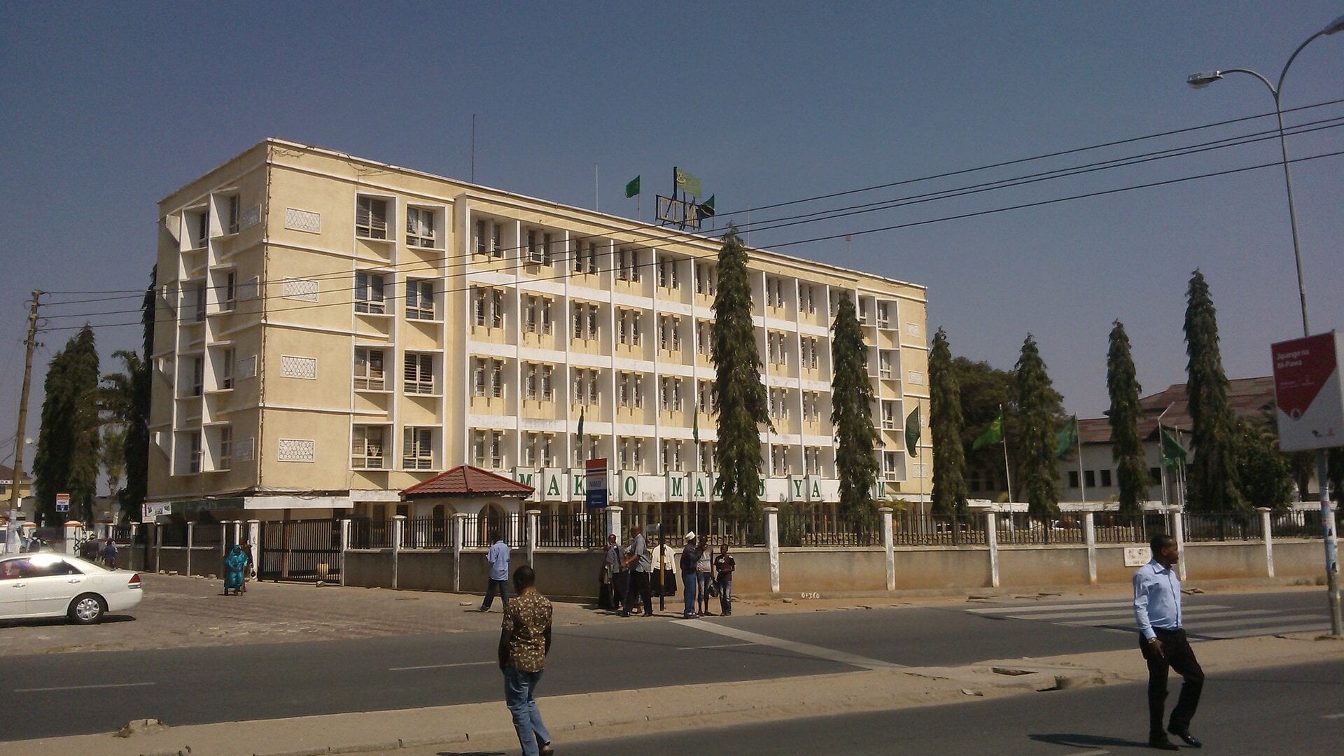 A five-storey cream building with the sign Makao Makuu ya CCM and green flags on its roof, with pedestrians in the street in front, in Dodoma, Tanzania