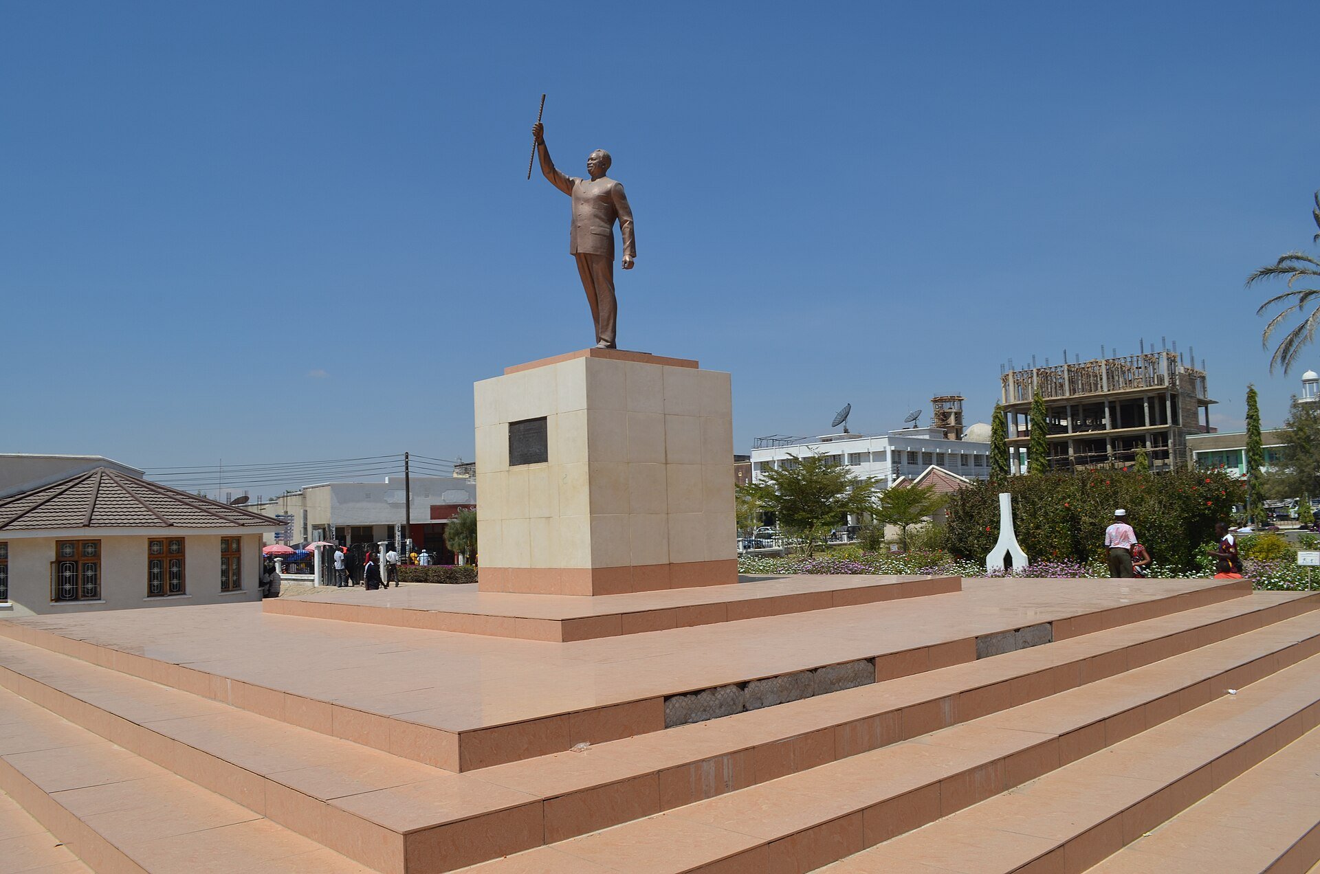 A bronze statue of Julius Nyerere with a raised arm on a stone plinth and wide steps in Nyerere Square, Dodoma, Tanzania, under a blue sky