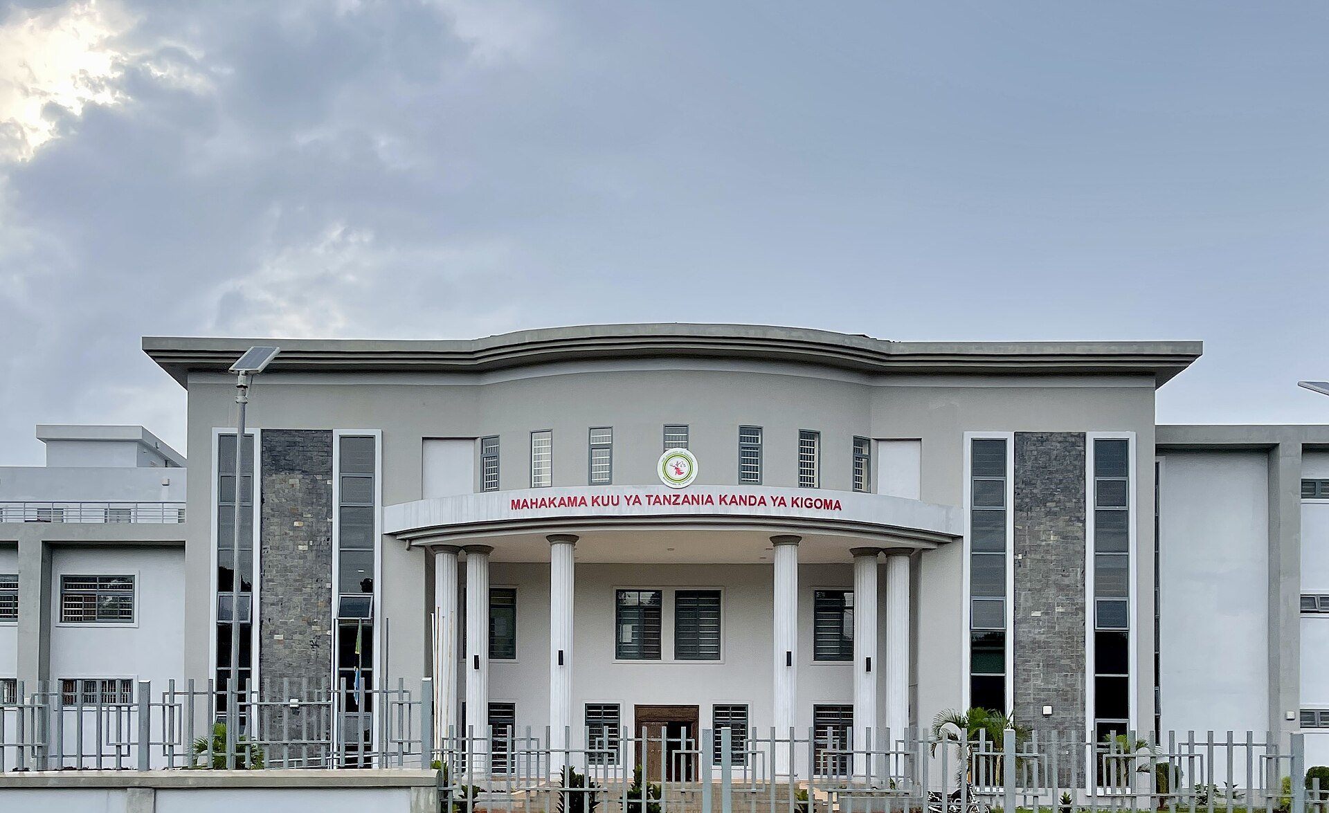The grey front of a court building with white columns and the red sign Mahakama Kuu ya Tanzania Kanda ya Kigoma under a cloudy sky in Kigoma, Tanzania