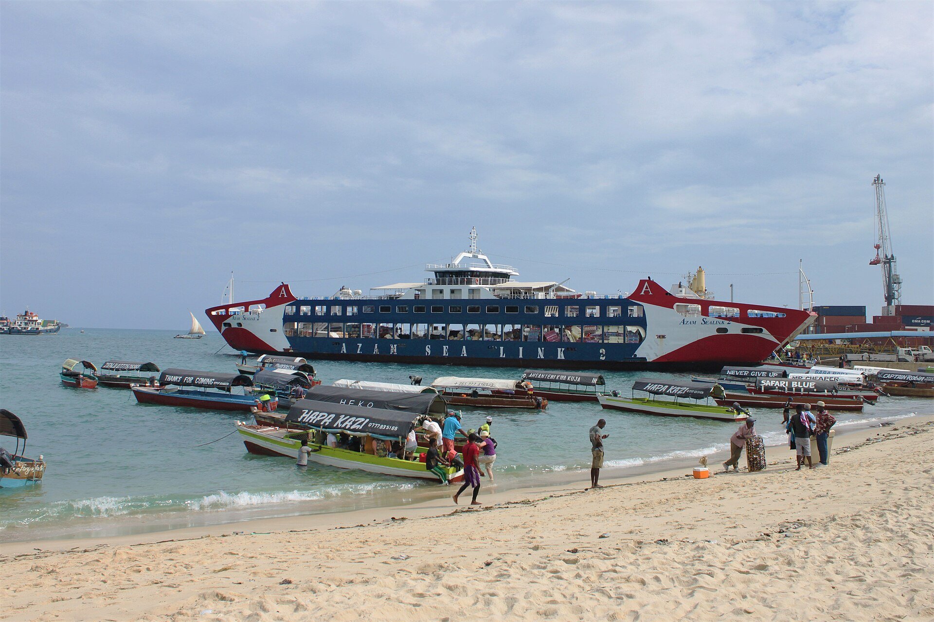 Ferry and small boats at the beach by the ferry terminal in Zanzibar City, with people on the sand and a container crane behind