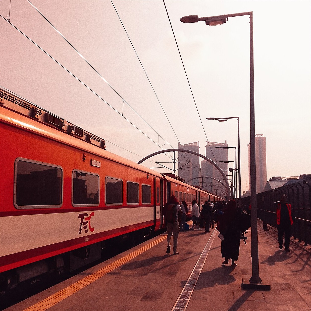 Passengers walk along a platform beside an orange-and-white electric passenger train under overhead power lines, with tower blocks in the background