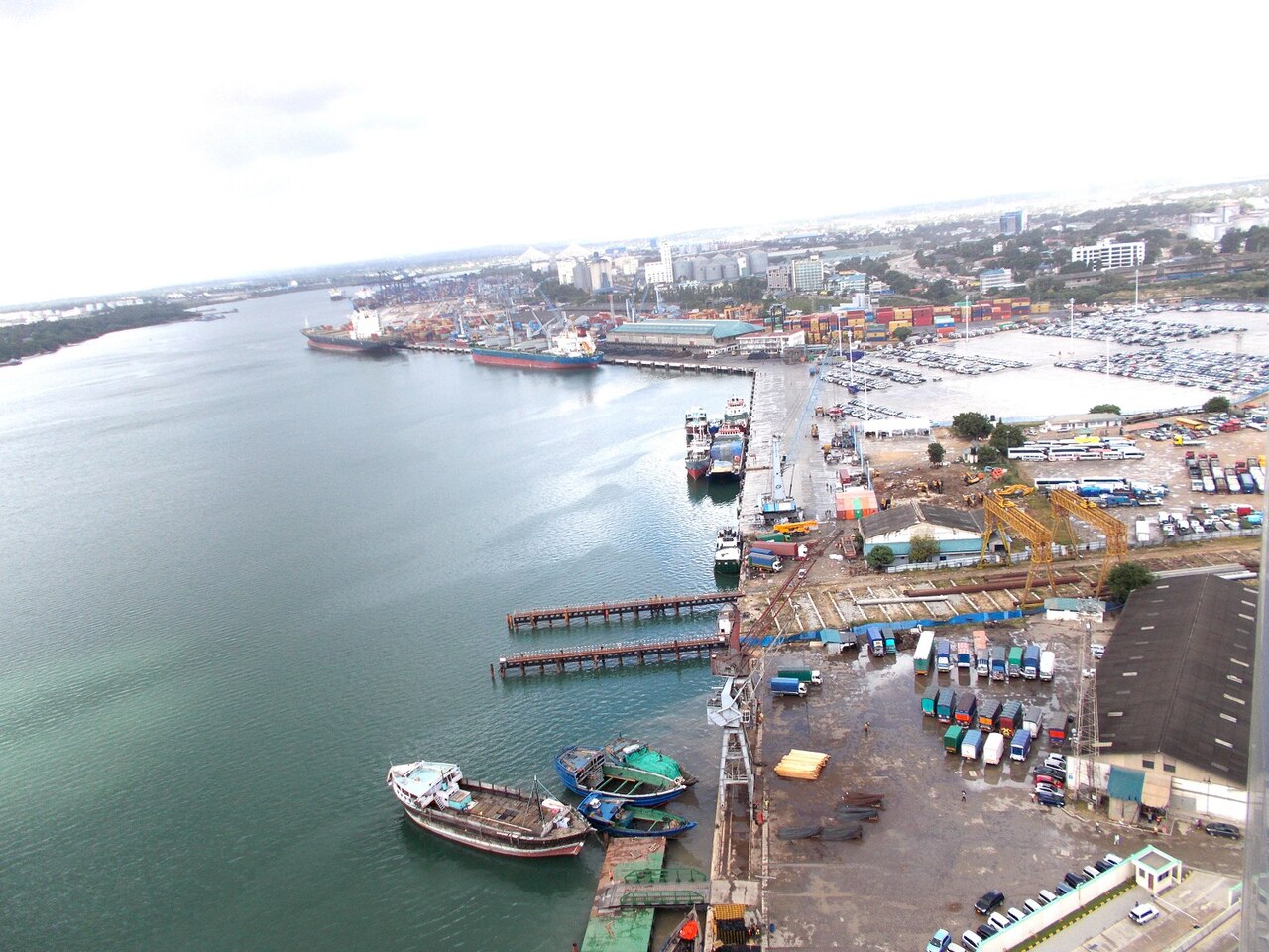 Aerial view of Dar es Salaam port with cargo ships at the quay, stacked containers, parked trucks and small wooden boats in the foreground