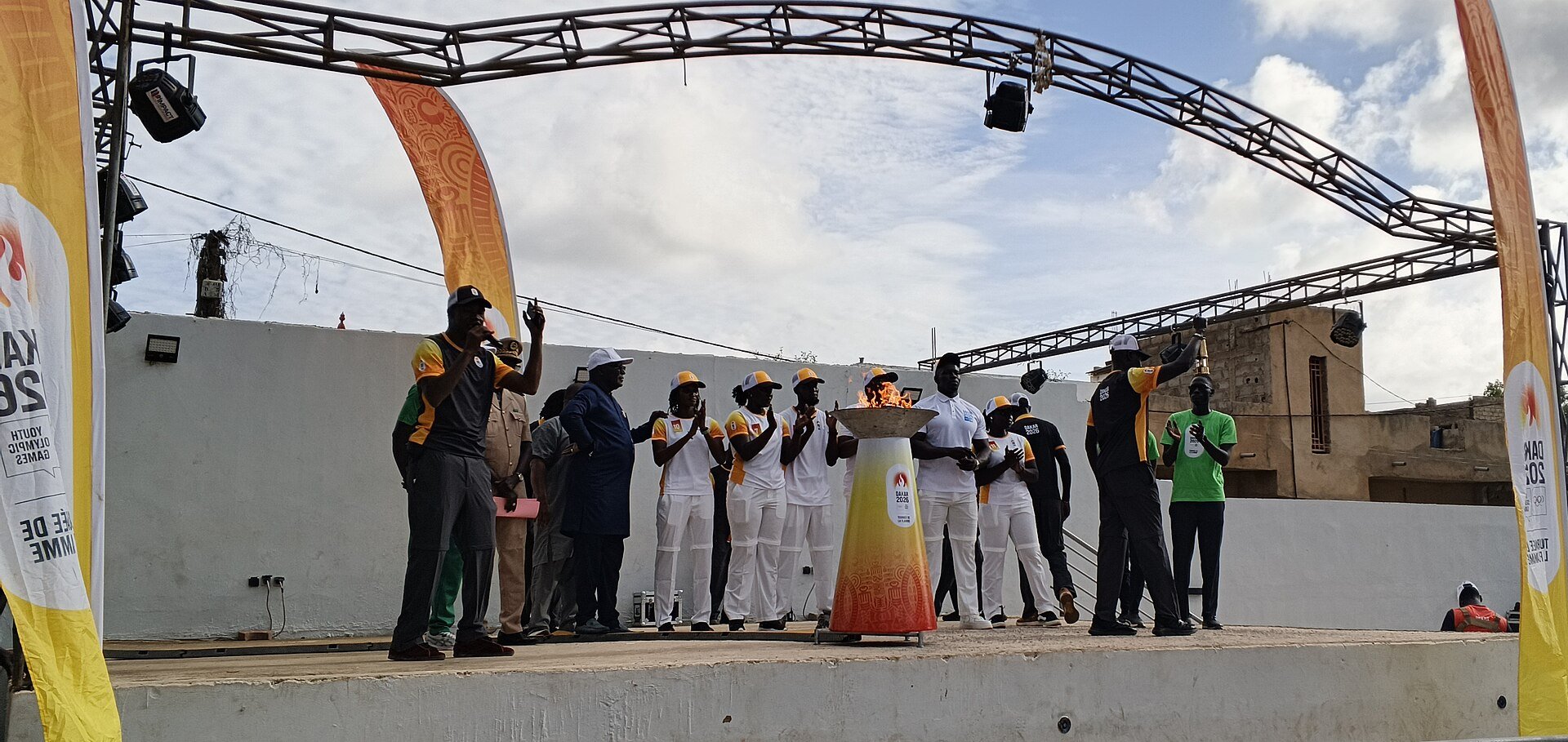 The Dakar 2026 flame cauldron burning on a stage during the Youth Olympic flame relay in Diamniadio with officials and torchbearers