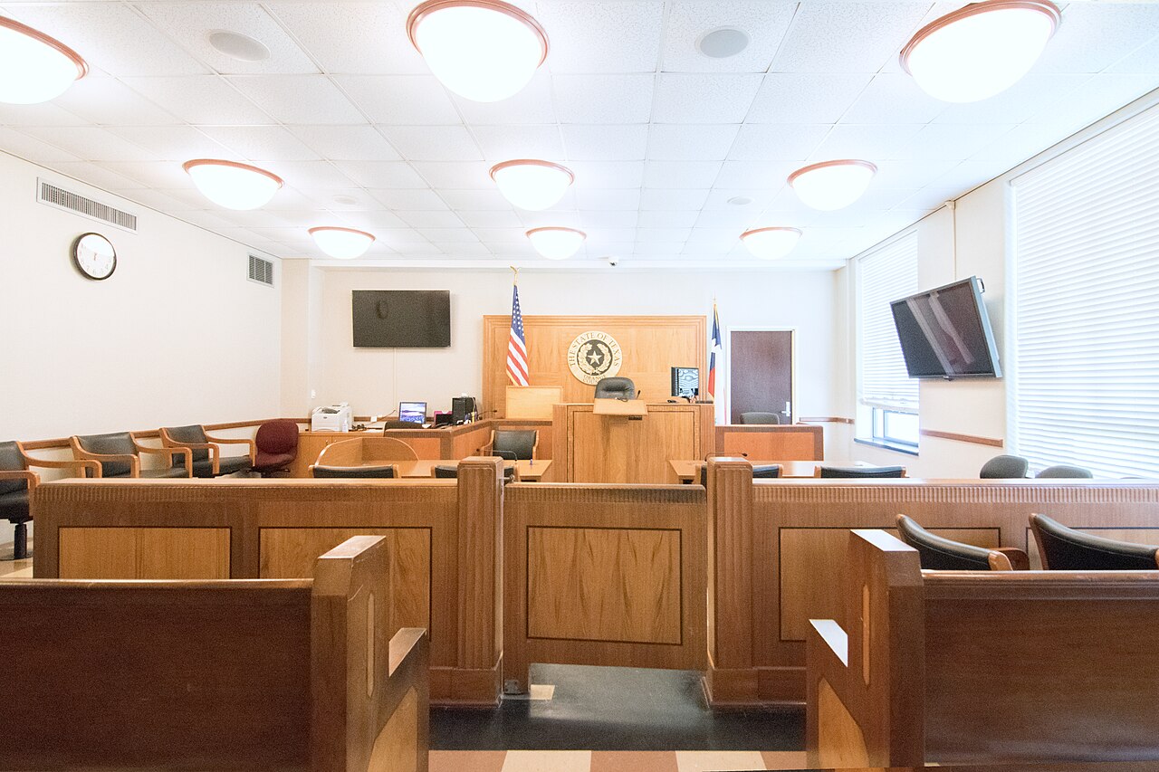 Empty courtroom with wooden benches and judge's bench, a State of Texas seal on the wall, and US and Texas flags