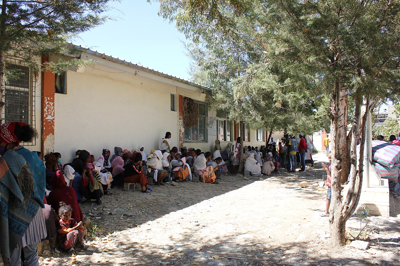 Displaced people sit and stand in the shade along a long building at a site in Shire, Tigray, Ethiopia
