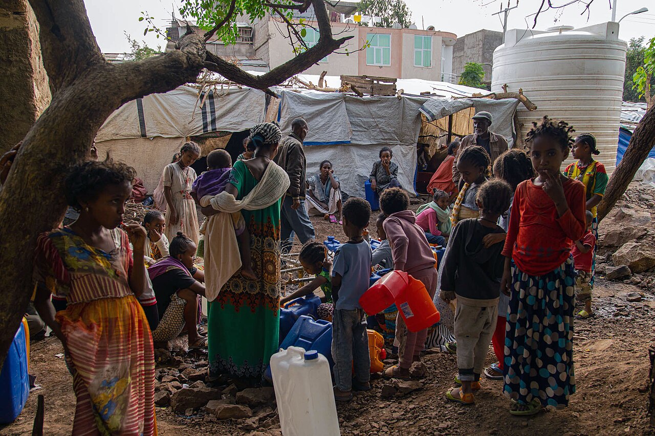 Displaced women and children fill jerrycans with water beside tarpaulin shelters and a large white water tank at a camp in Shire, Tigray