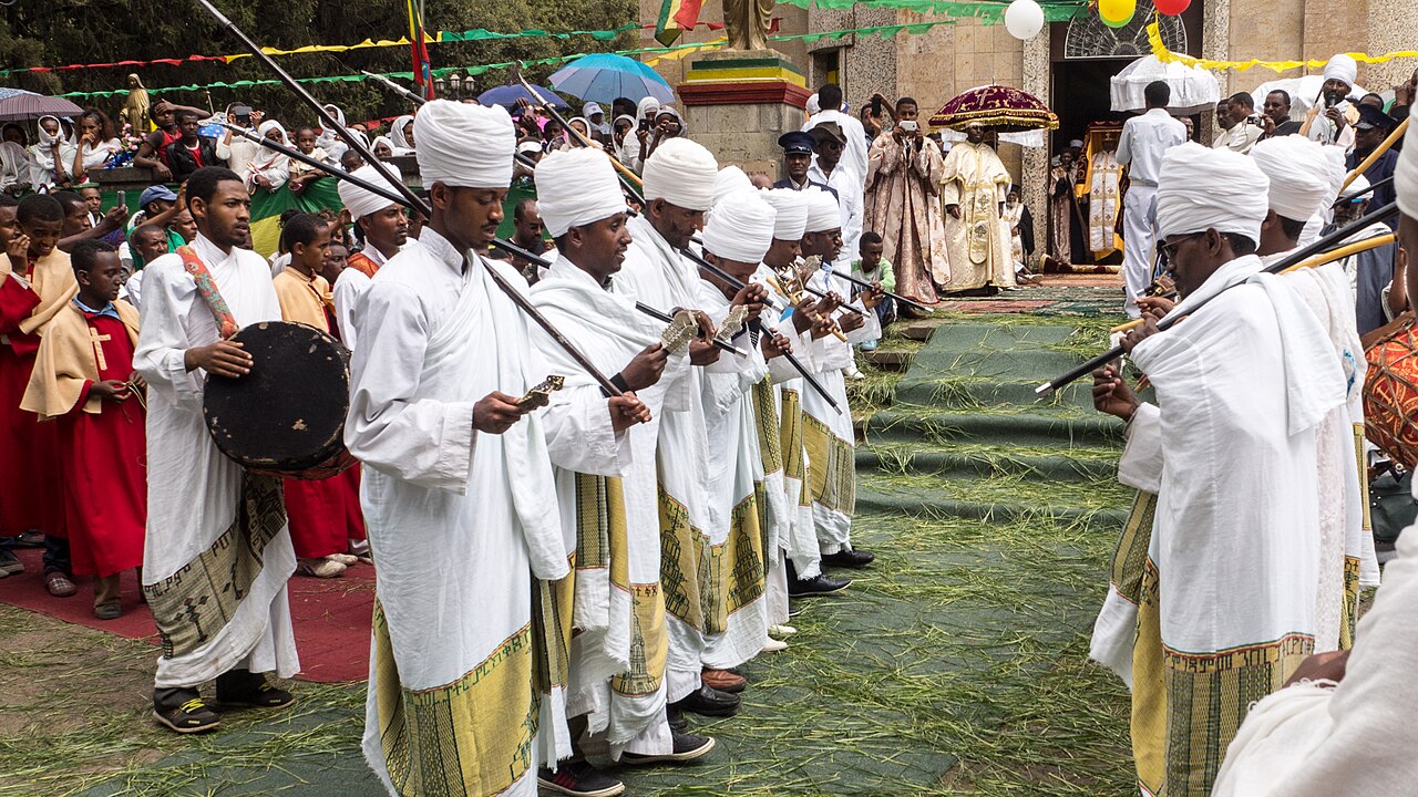 Orthodox priests in white robes with prayer sticks and a drum stand in two lines on a grass-strewn carpet outside a church during Timkat in Addis Ababa.