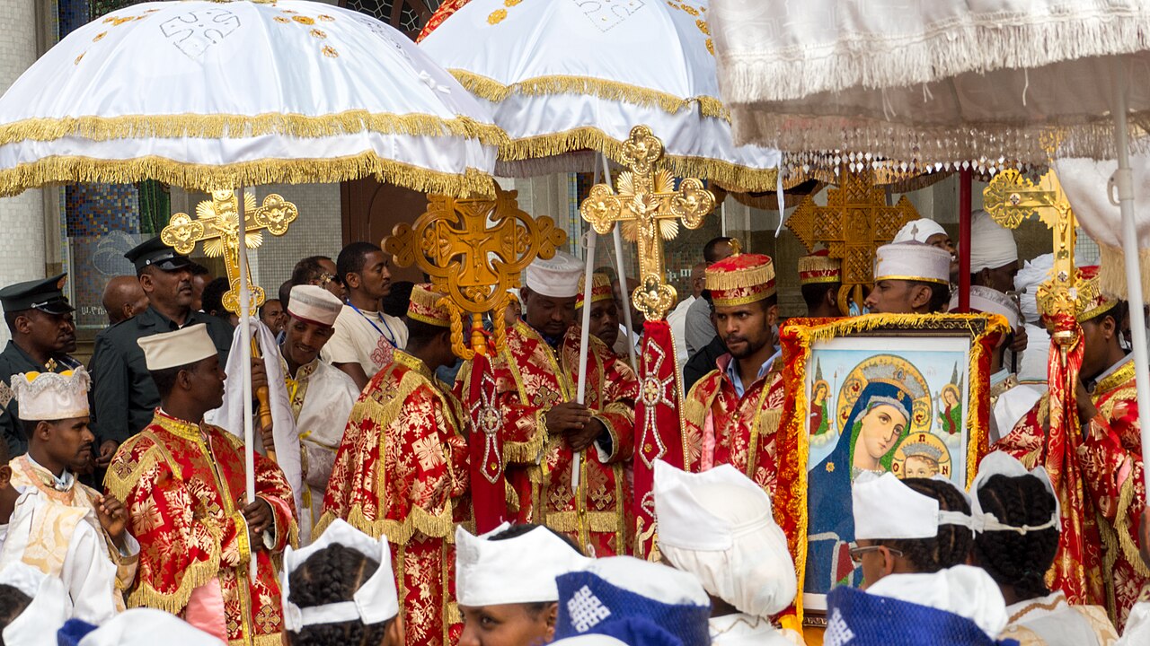 Priests in red and gold robes stand under white umbrellas with gold crosses beside a framed painting of the Virgin Mary during Timkat in Addis Ababa.