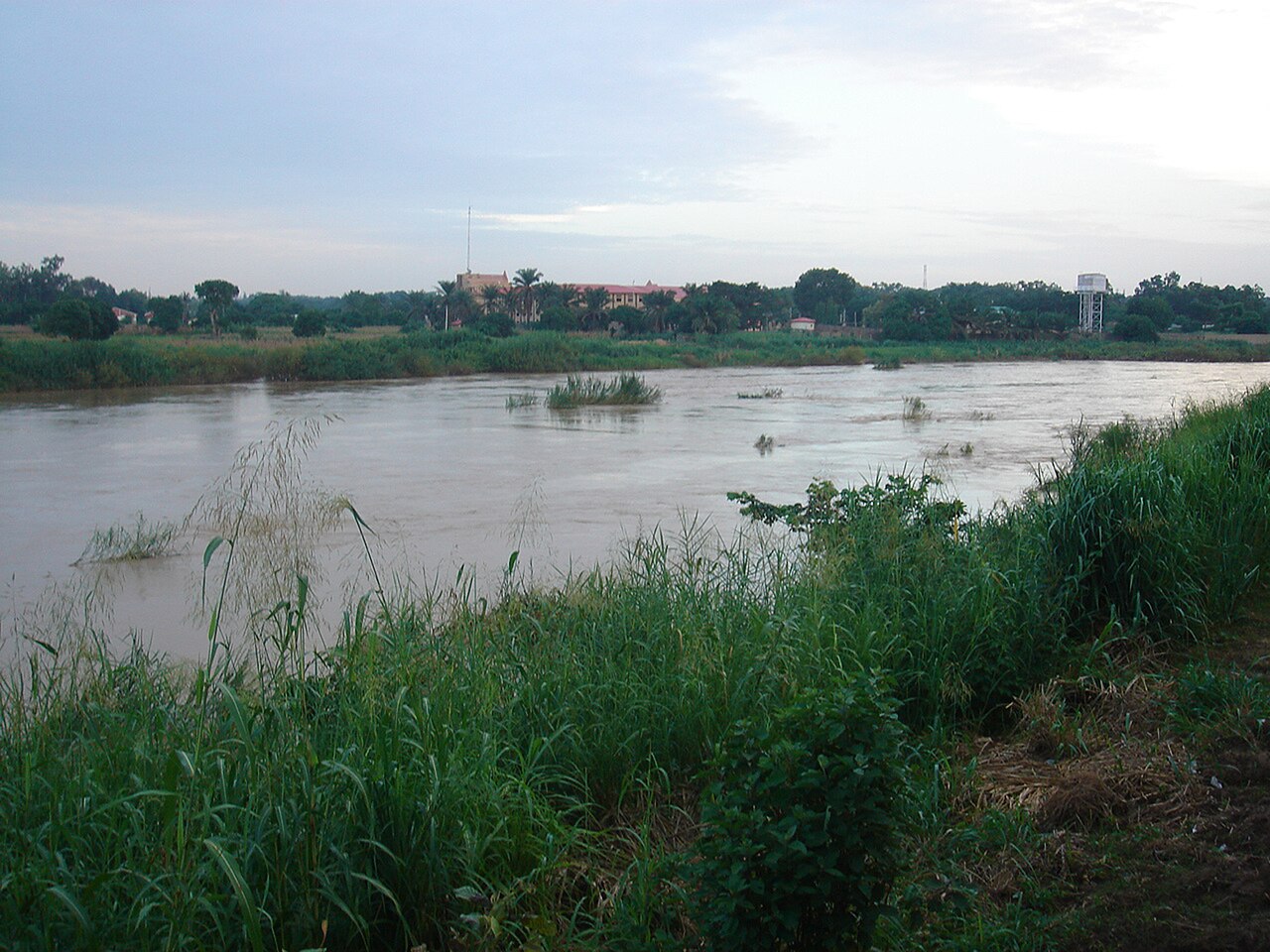 The Kaduna River flows brown between green grassy banks, with trees, a building and a water tower on the far shore under a pale sky