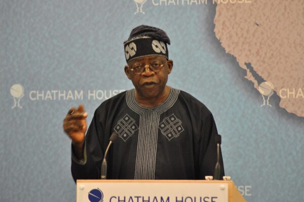 Bola Tinubu in a black embroidered agbada and cap speaks at a lectern with a Chatham House backdrop and a map of West Africa behind him
