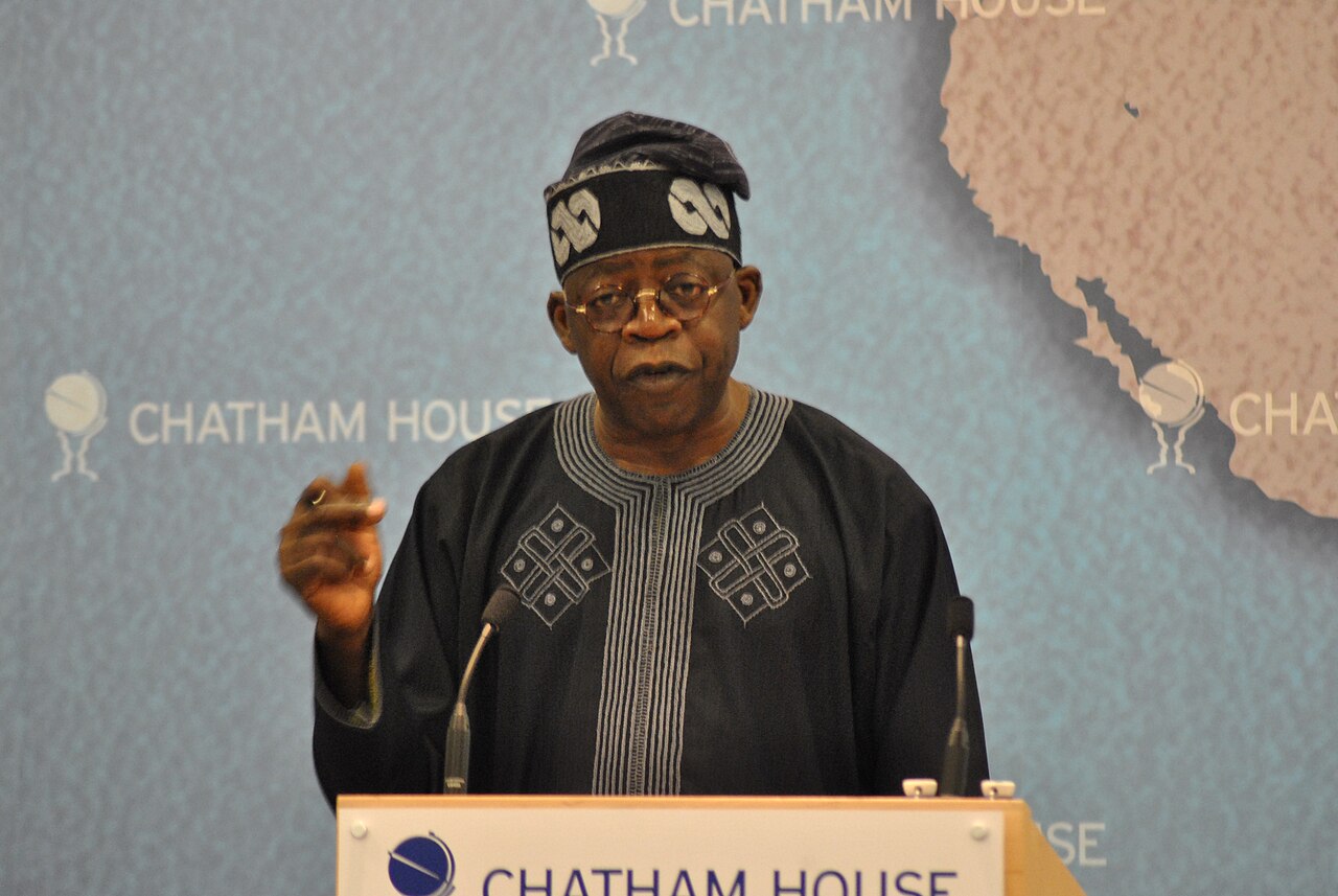 Bola Tinubu in a black embroidered agbada and cap speaks at a lectern with a Chatham House backdrop and a map of West Africa behind him