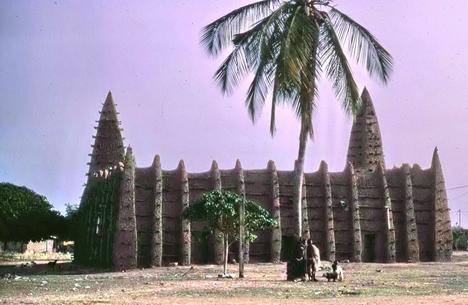 The mud-walled mosque of Kong in northern Ivory Coast, with a palm tree and people in front