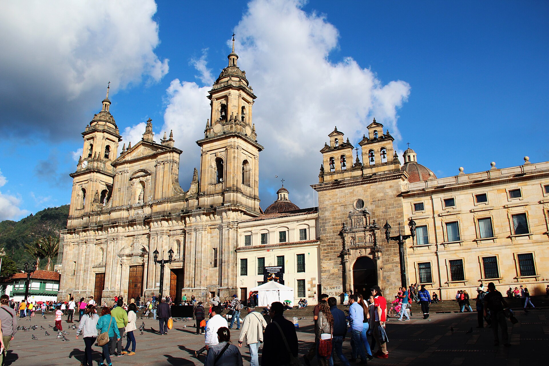 The twin-towered Primatial Cathedral on Bolívar Square in Bogotá under a blue sky with white clouds, with pedestrians and pigeons in the square