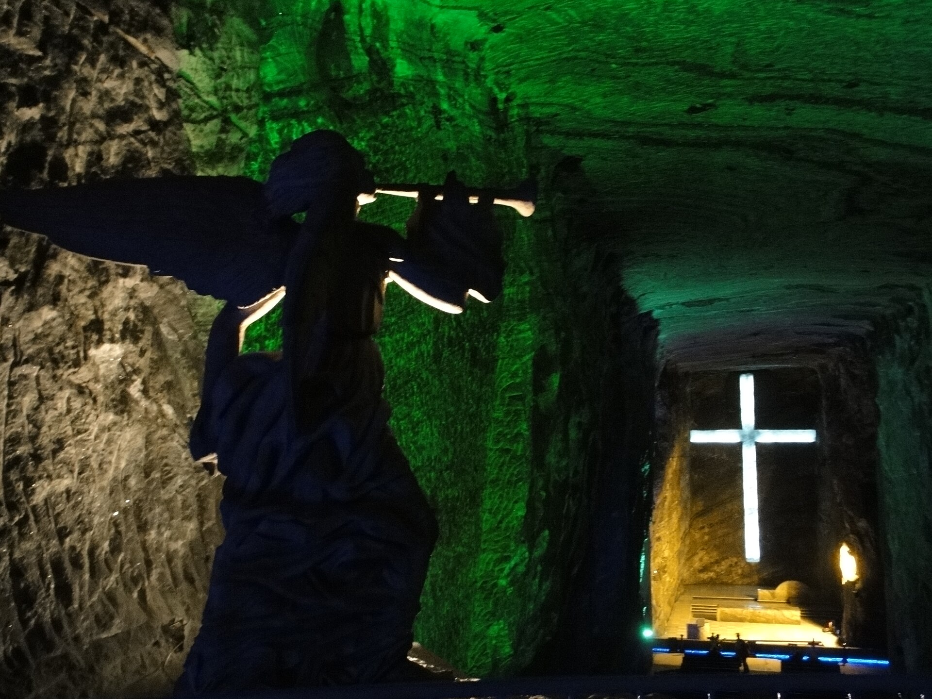 Inside the Zipaquirá Salt Cathedral, a dark silhouette of a winged angel blowing a trumpet in the foreground and a lit white cross at the end of a green-lit salt tunnel