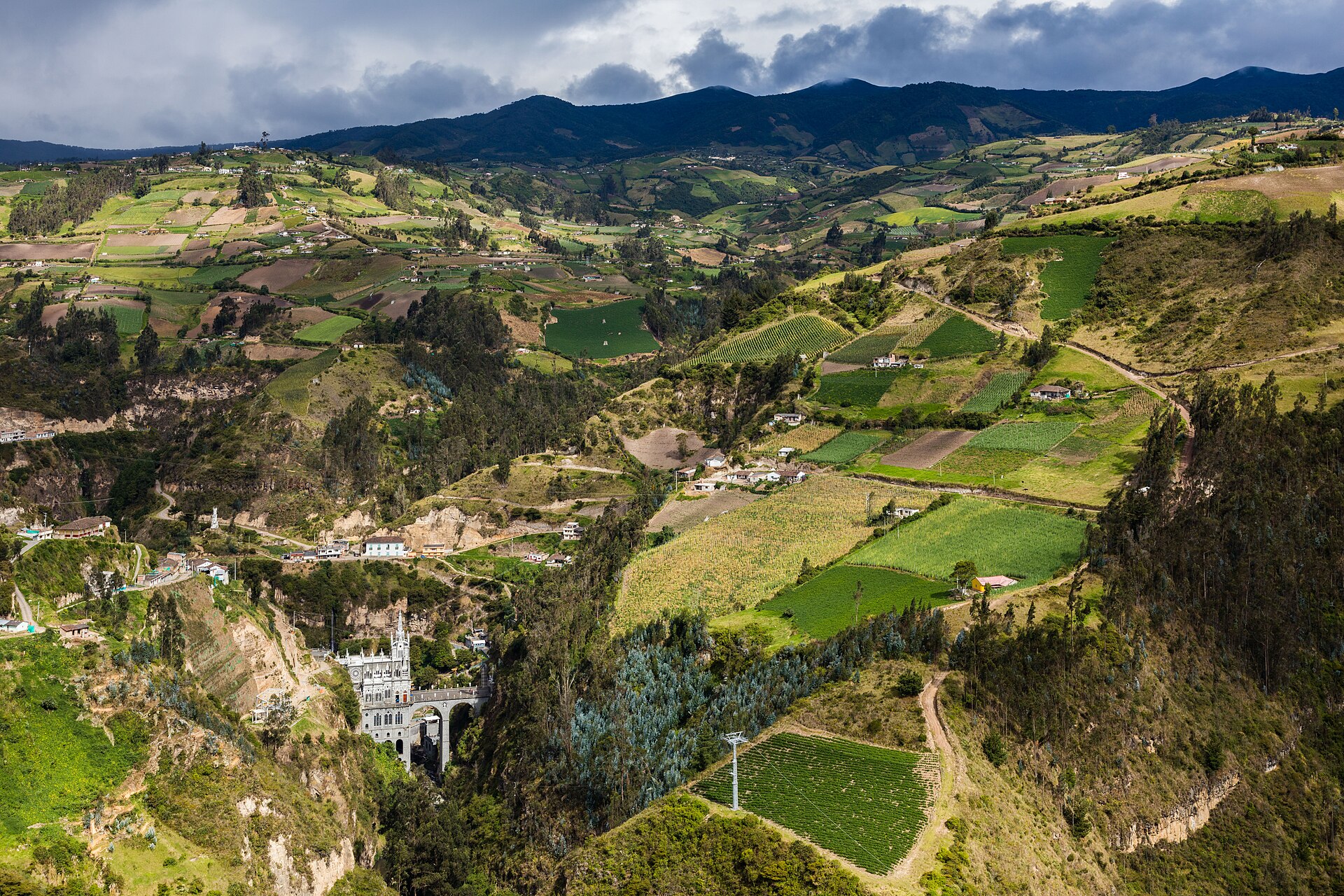 The Las Lajas Sanctuary, a white church built across a deep canyon, seen from a distance among green farmland and mountains near Ipiales