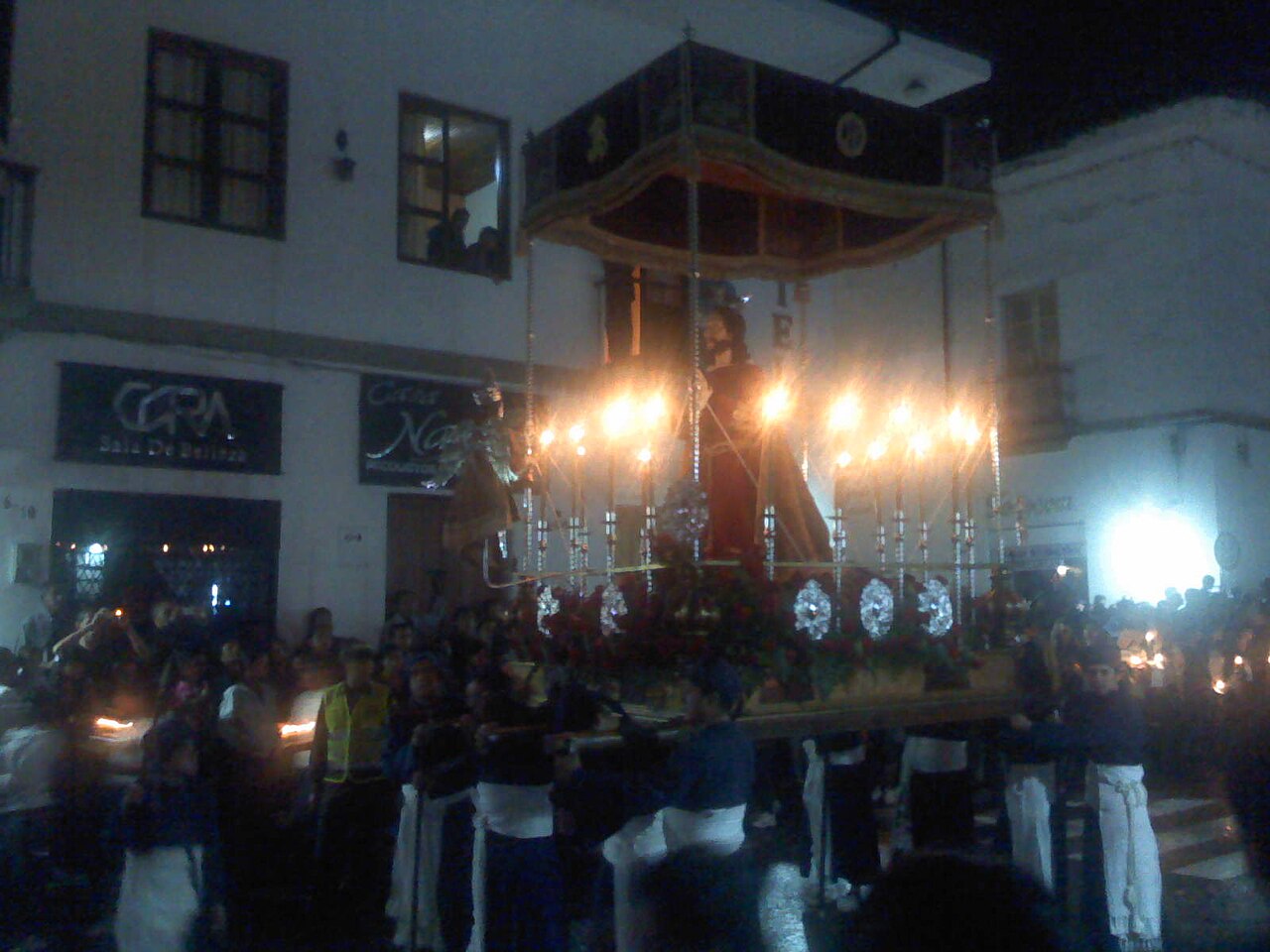 A night Holy Week procession in Popayán: bearers in blue and white carry a candle-lit float with a canopy and a statue through a street lined with spectators
