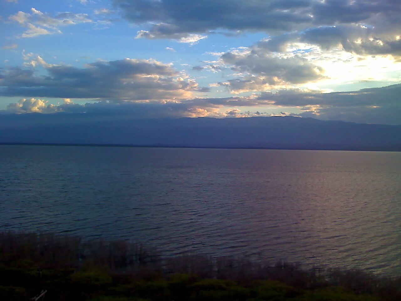 Calm water of Lago Enriquillo at dusk under a cloudy sky with mountains on the far shore