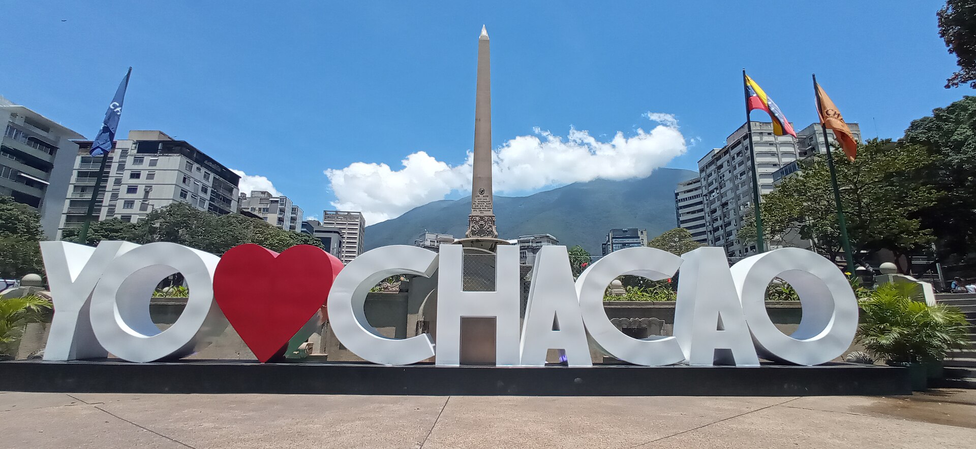 The Altamira obelisk at Plaza Francia in Caracas behind a large 'Yo amo Chacao' sign, with the Avila mountain and a blue sky beyond