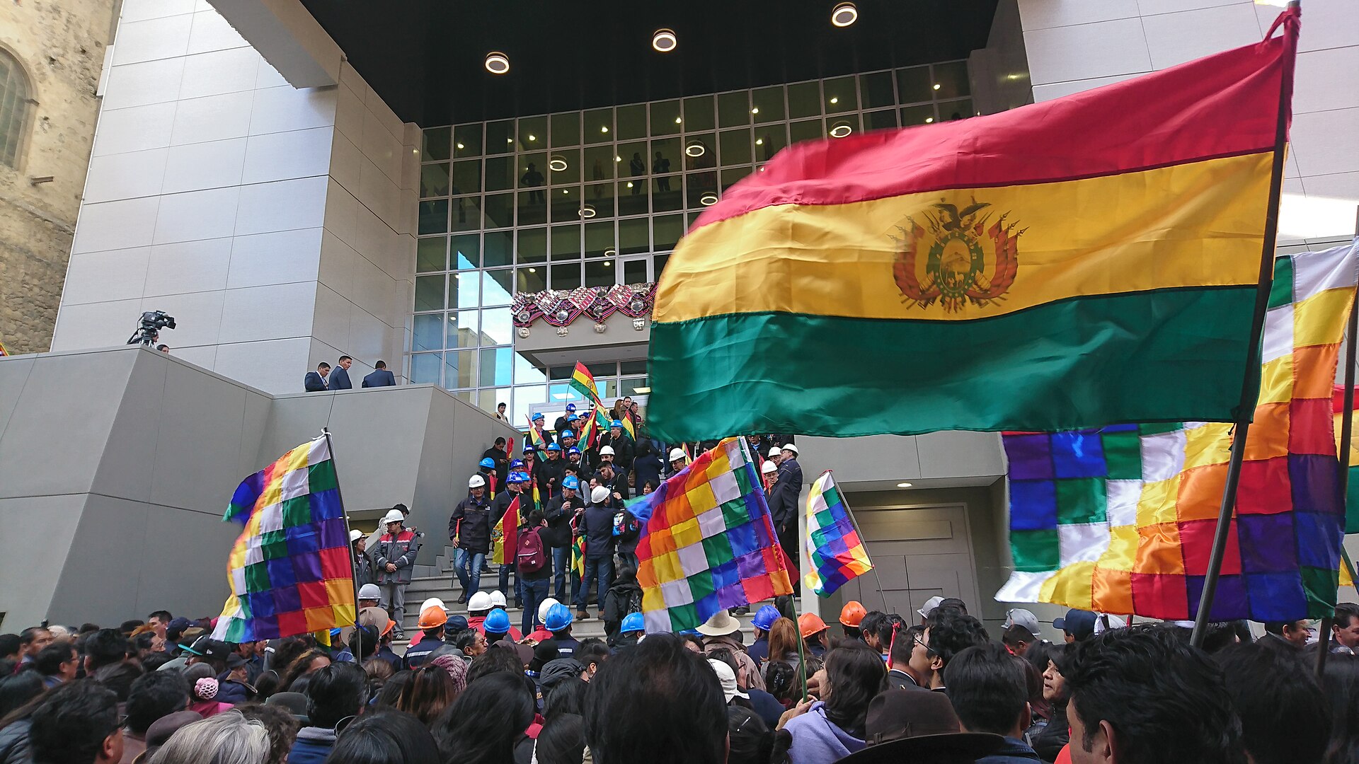 A crowd carrying Bolivian flags and wiphala banners outside the glass entrance of the Casa Grande del Pueblo in La Paz