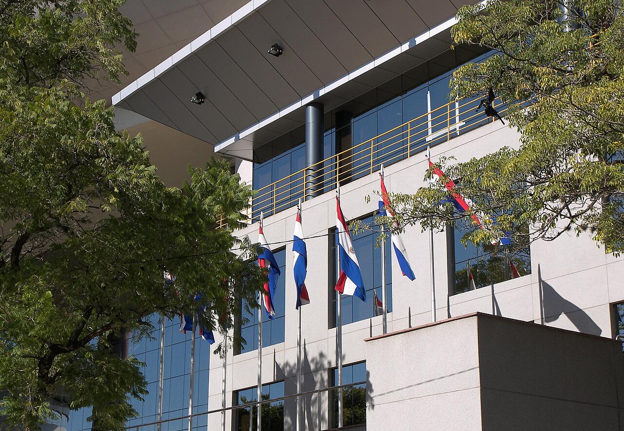 A glass-fronted Congress building in central Asunción with Paraguayan flags flying below its entrance canopy, framed by trees
