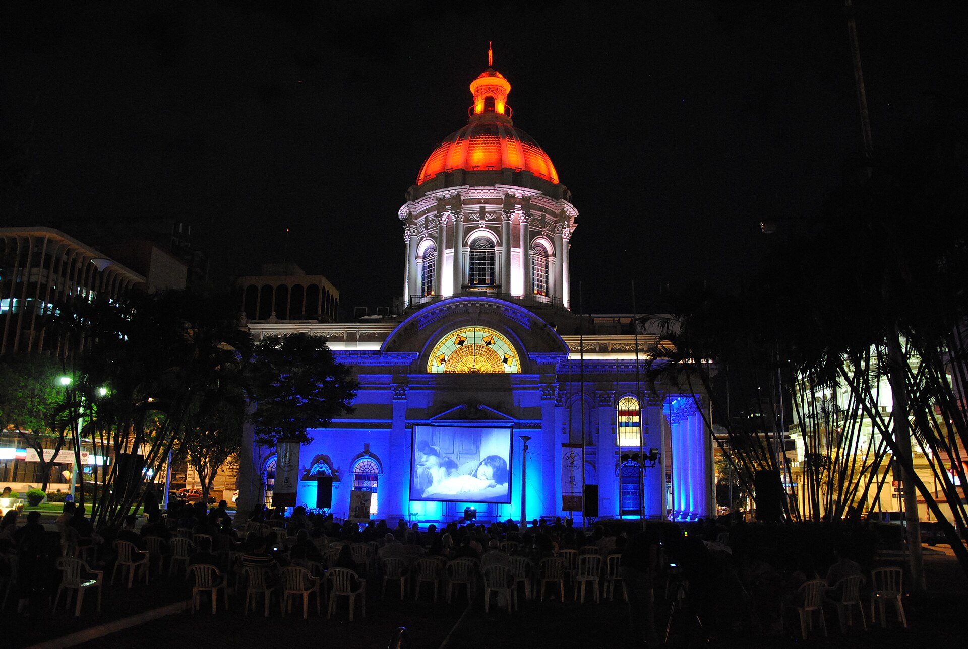 The domed National Pantheon of the Heroes in Asunción lit in blue and orange at night, with an audience watching an outdoor film screening