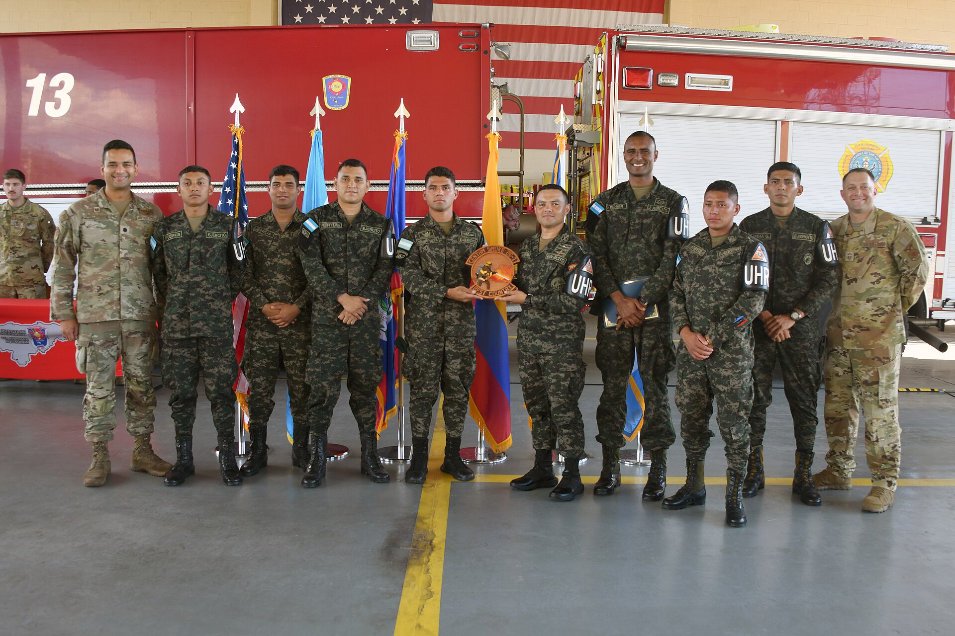 US service members and a row of Honduran personnel in camouflage pose in front of a red fire engine, with US, Honduran and other flags