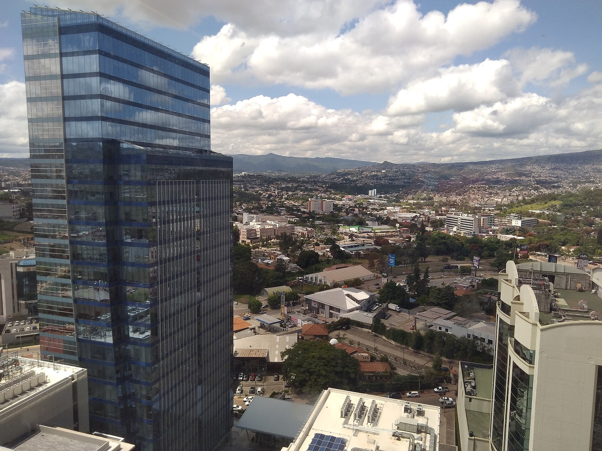 Glass office towers and the hills of Tegucigalpa under a cloudy sky, seen from a high floor