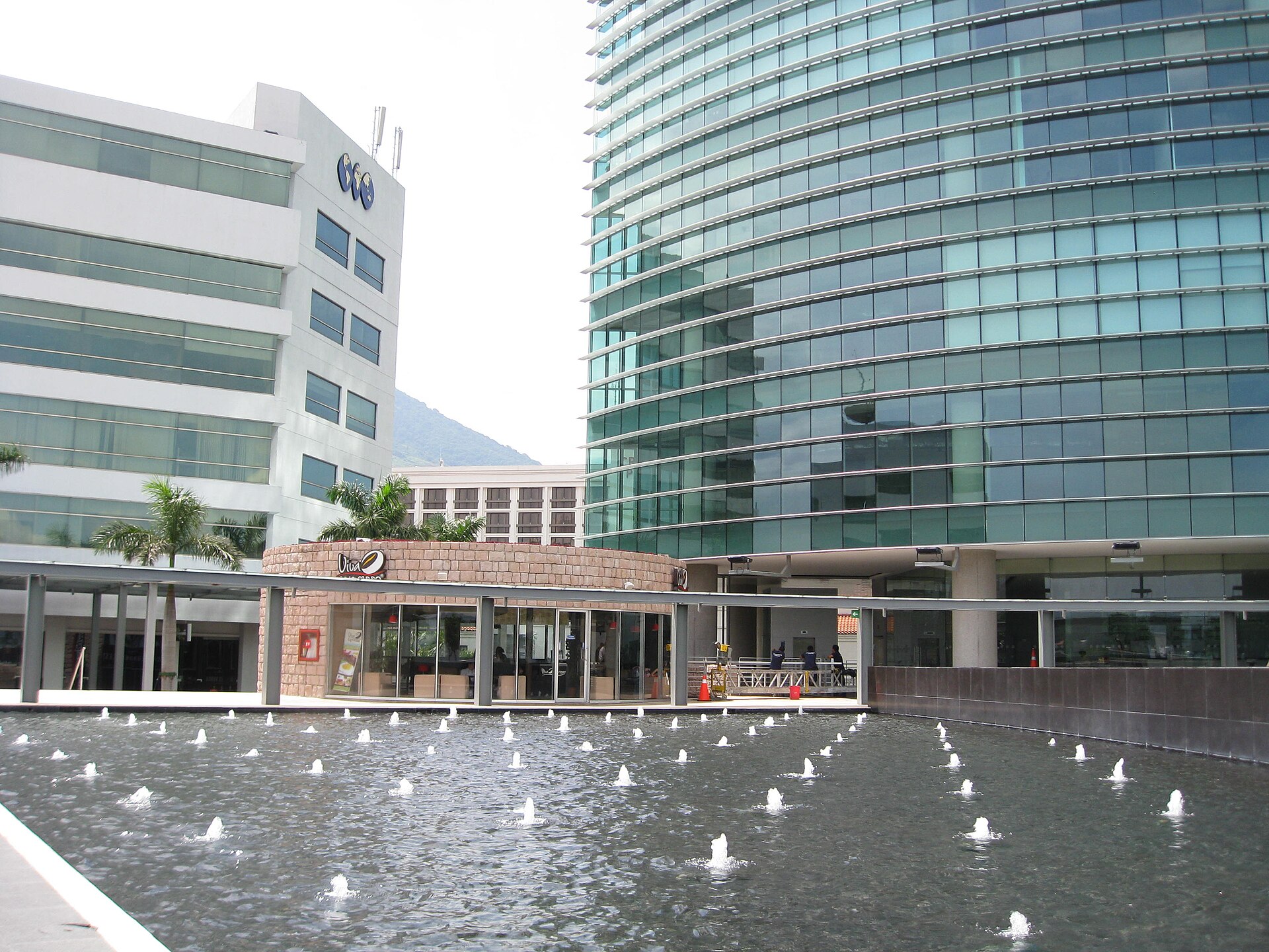 A fountain pool in front of glass office towers at Plaza Futura in San Salvador, with a green hill behind