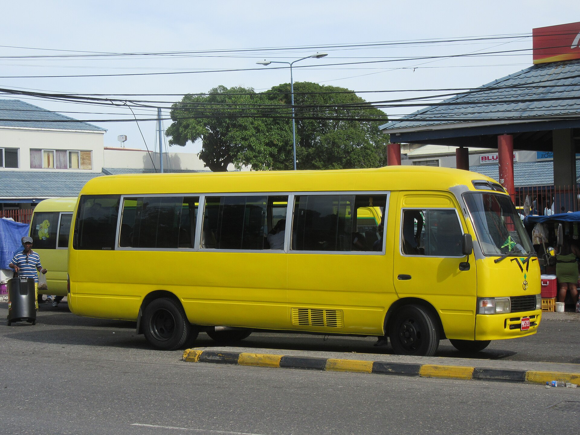 A yellow Toyota Coaster minibus stopped at a kerb beside a covered terminal at Half Way Tree, Kingston