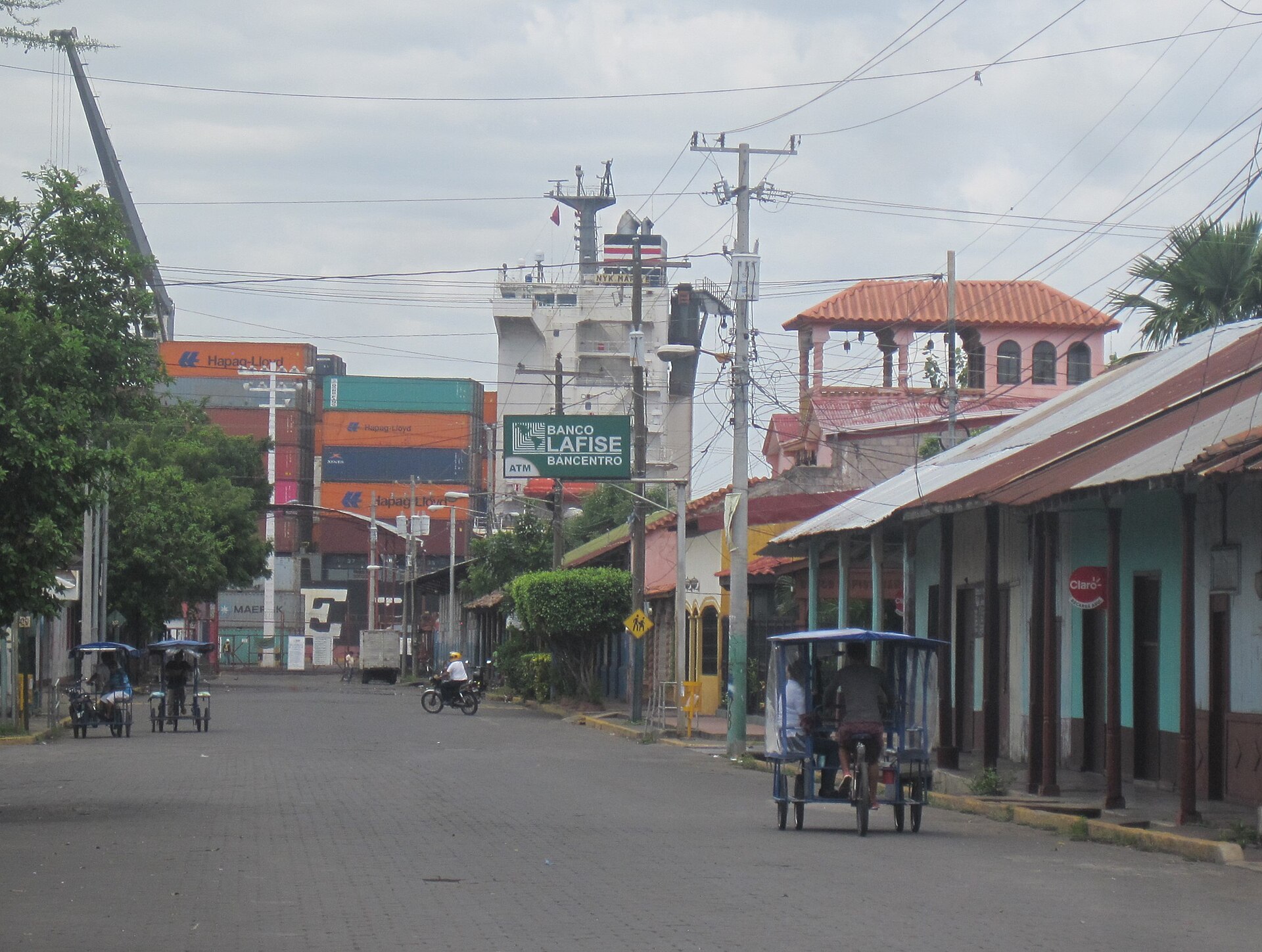 A street in Corinto, Nicaragua, with stacked shipping containers and a cargo ship behind low buildings and bicycle taxis