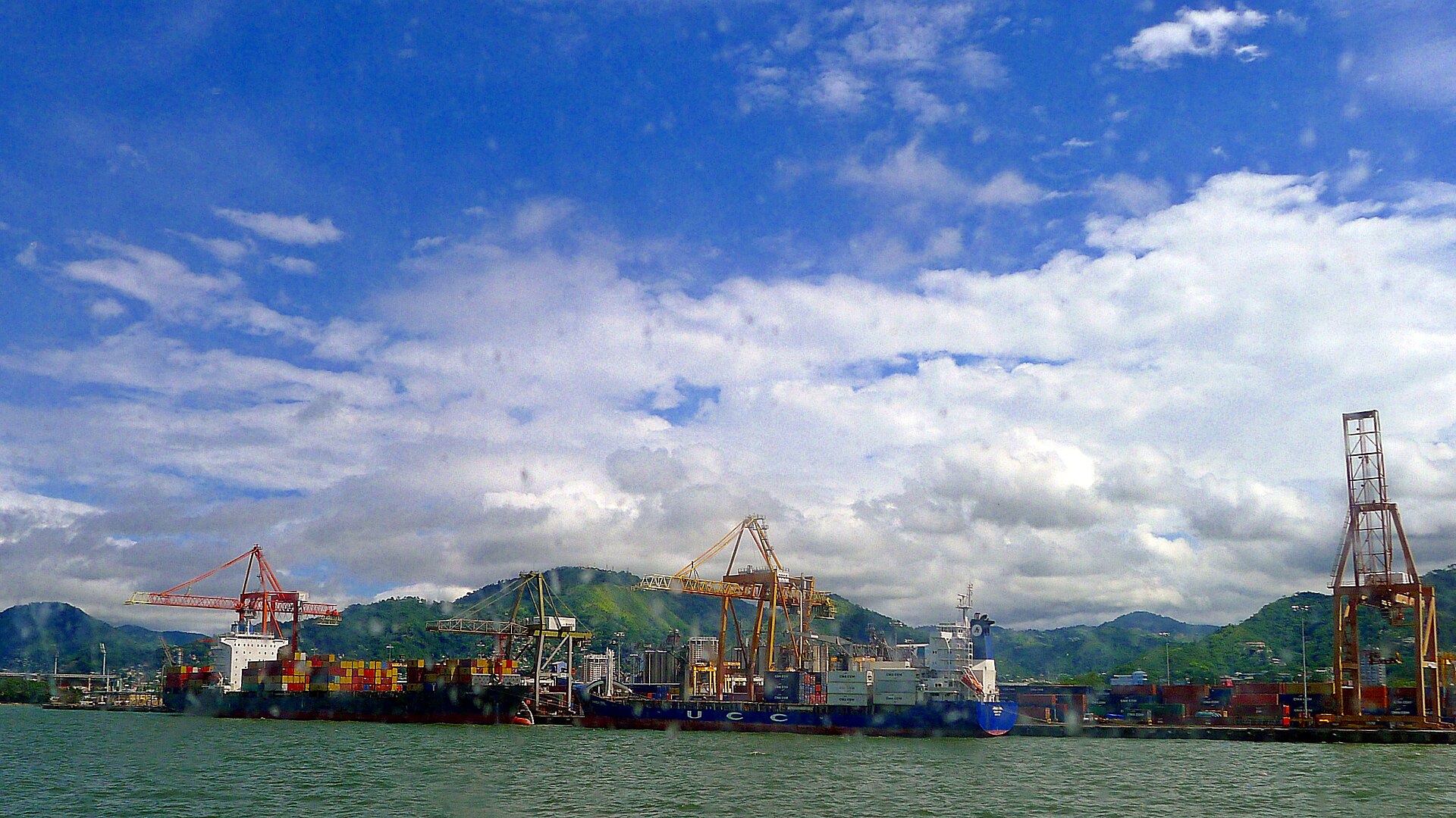 Container ships and orange cranes at a port under a blue sky, with green hills behind