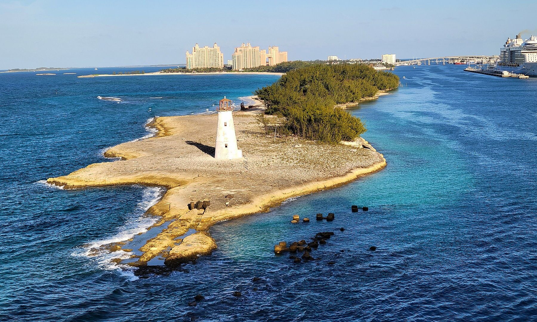 A white lighthouse on a rocky, scrub-covered point at the mouth of Nassau harbour, with resort towers behind and a cruise ship at right