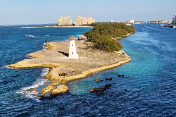 A white lighthouse on a rocky, scrub-covered point at the mouth of Nassau harbour, with resort towers behind and a cruise ship at right