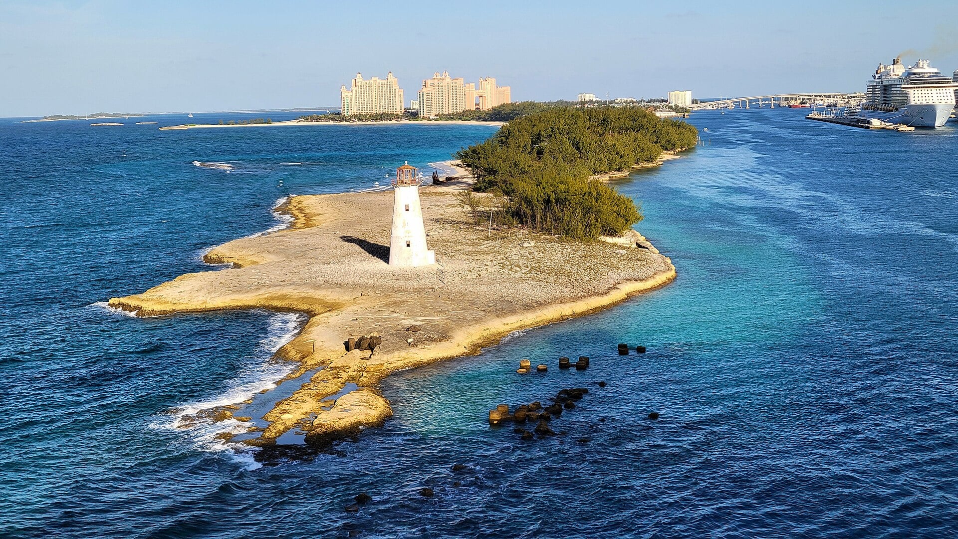 A white lighthouse on a rocky, scrub-covered point at the mouth of Nassau harbour, with resort towers behind and a cruise ship at right