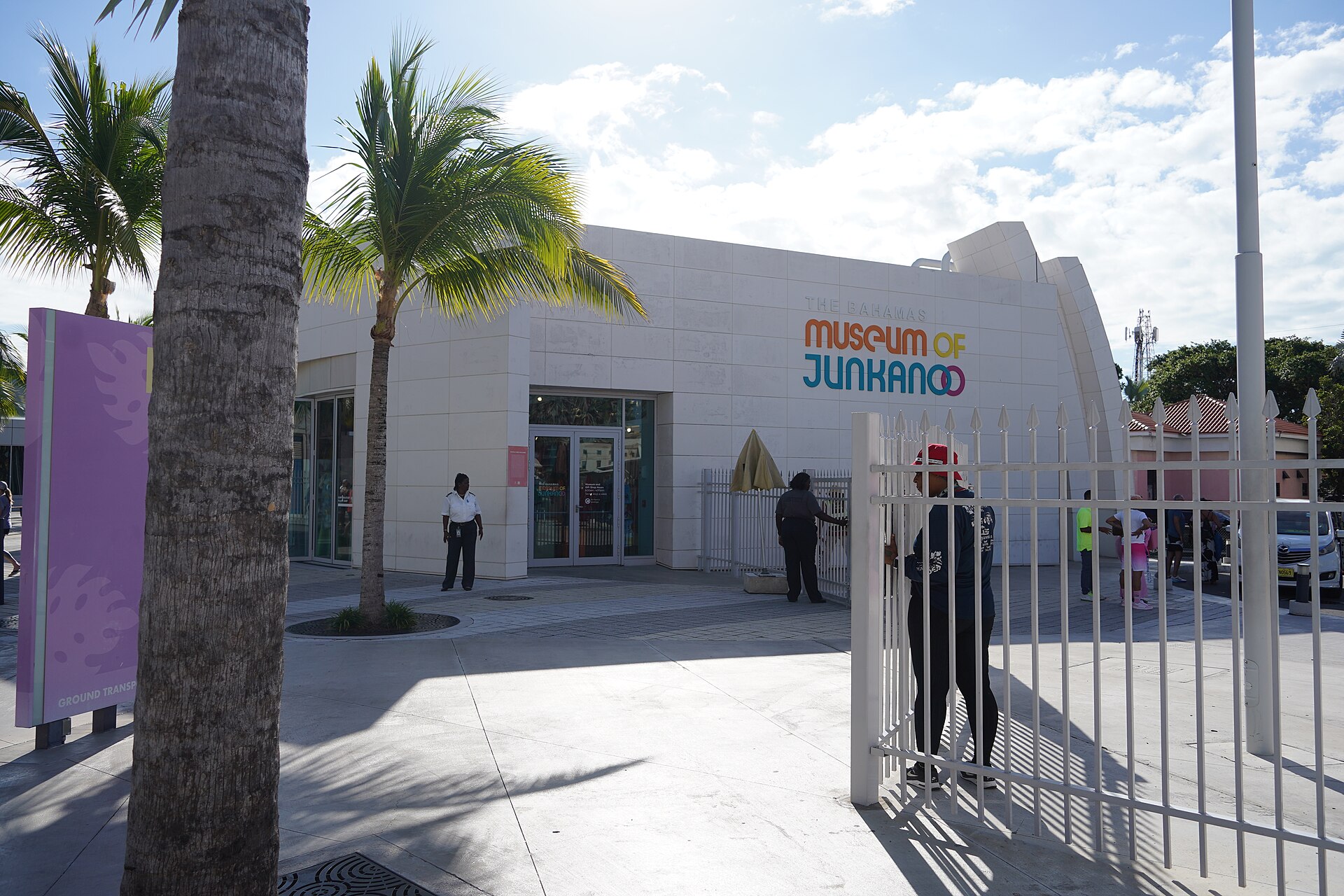 A white modern building with the sign Museum of Junkanoo, palm trees in front and visitors at its gate