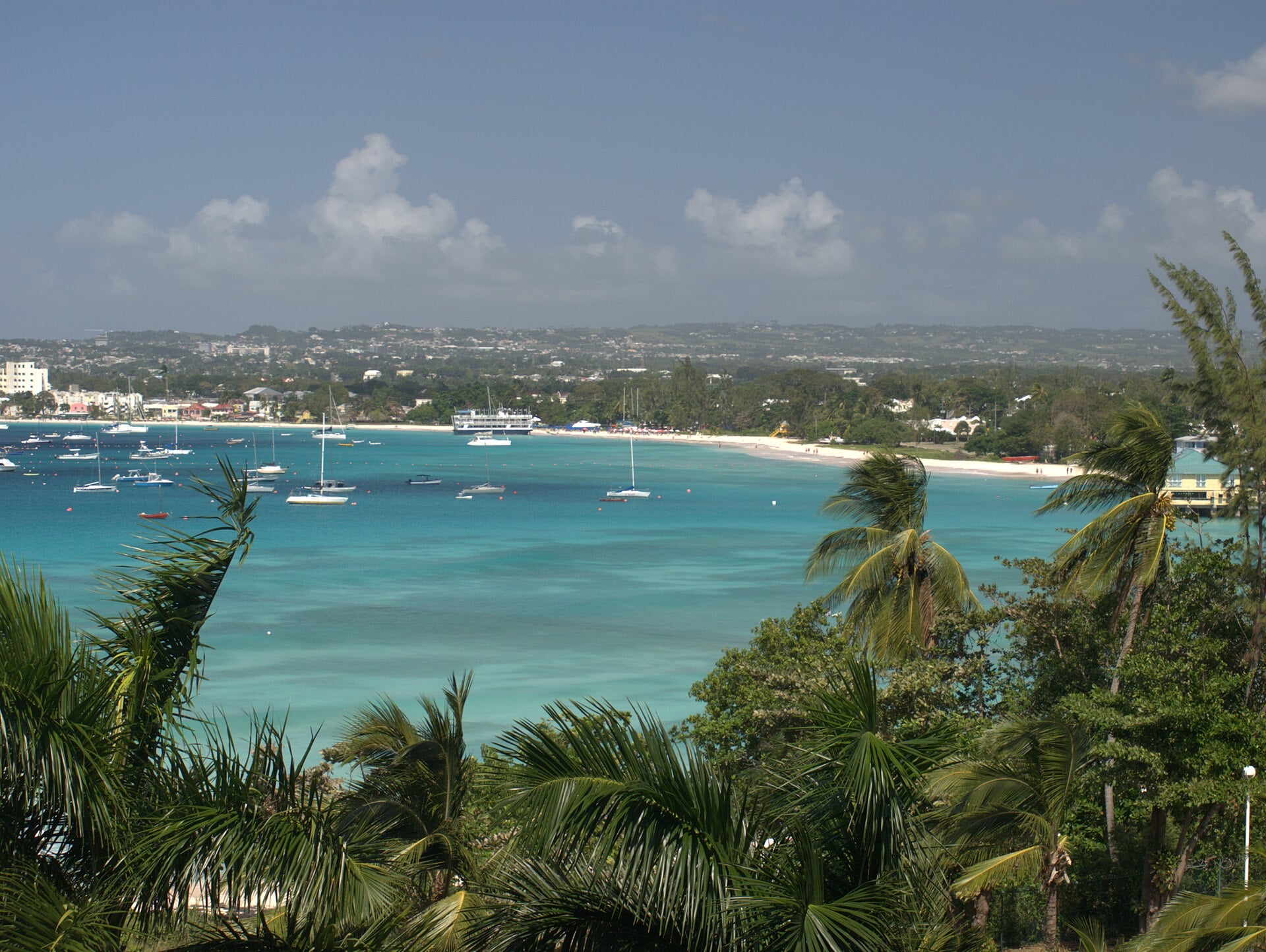 Turquoise Carlisle Bay with moored boats, a white beach and the Bridgetown coastline seen through palm trees