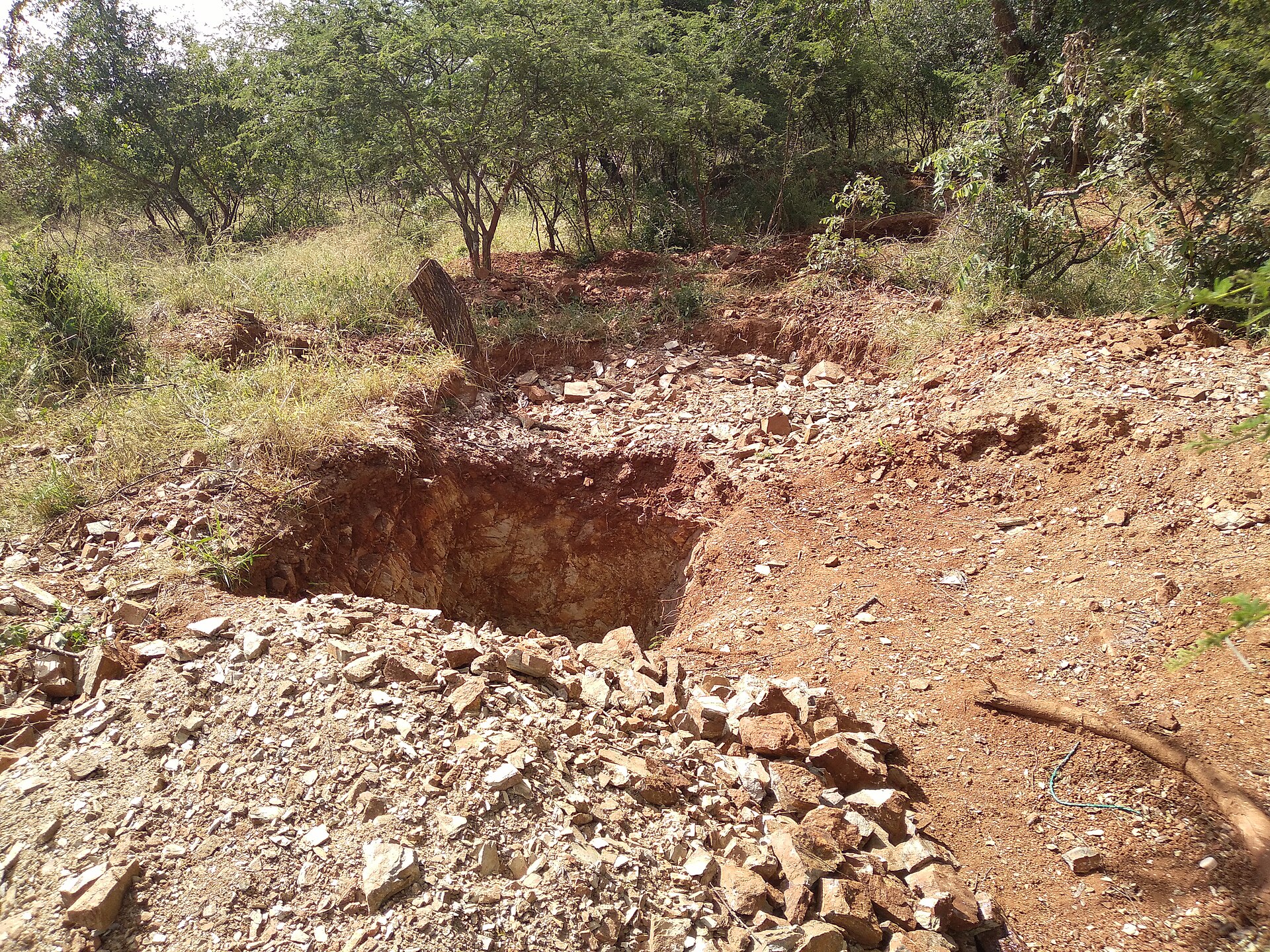 A shallow pit dug into red soil, with piles of broken rock beside it and bush behind