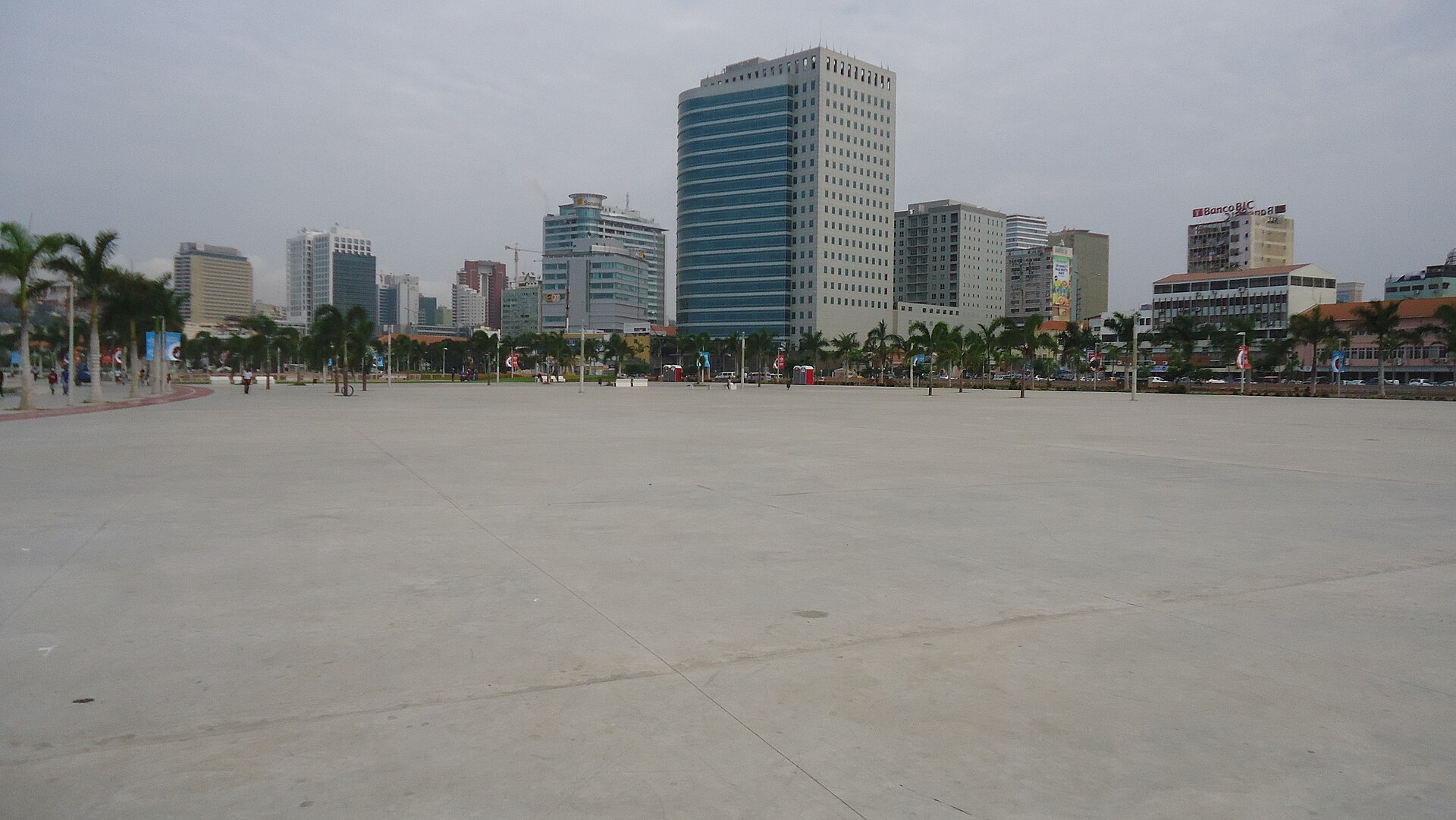 A wide paved square beside Luanda's Marginal avenue with palm trees and office towers, one with a bank sign, in the background