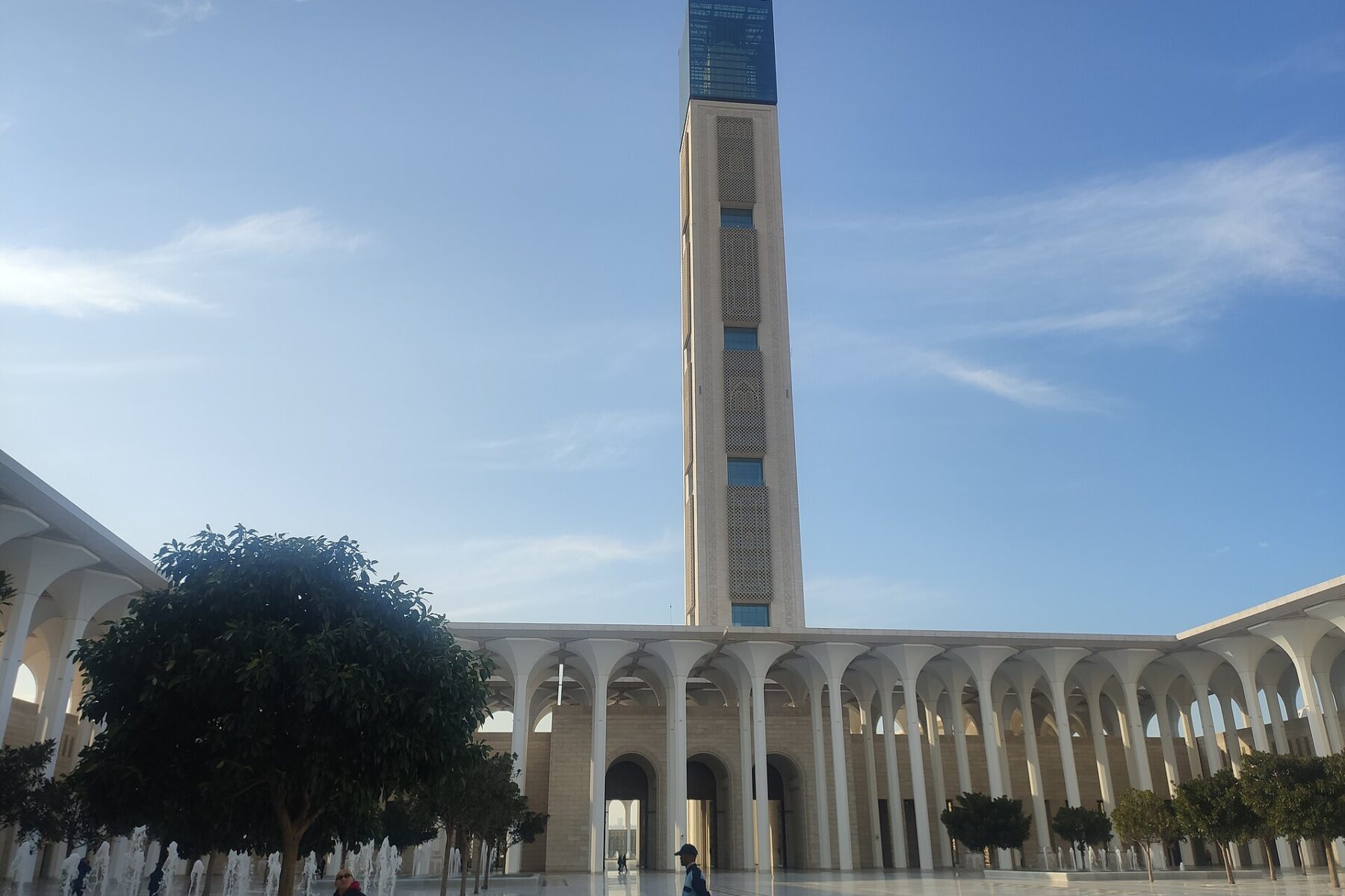 The tall minaret and white arcades of the Great Mosque of Algiers rising over a fountain courtyard under a blue sky, with a few visitors in the foreground