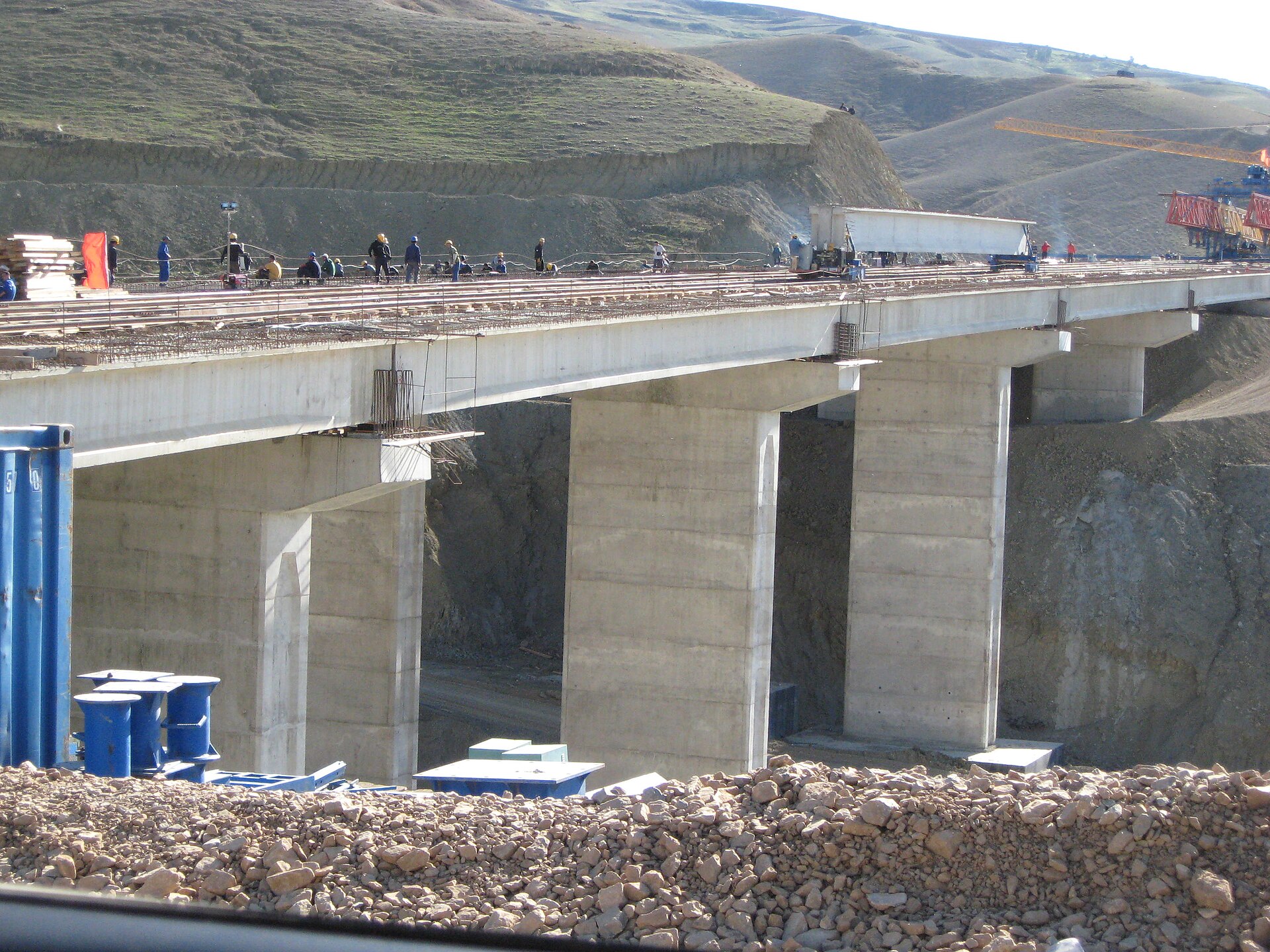 A concrete motorway viaduct on two pairs of pillars under construction across a dry valley, with workers on the deck and a crane on the hillside